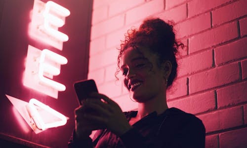 Frau steht vor einer Steinwand mit einem pinken Neonschild und scrollt am Smartphone Frau steht vor einer Steinwand mit einem pinken Neonschild und scrollt am Smartphone