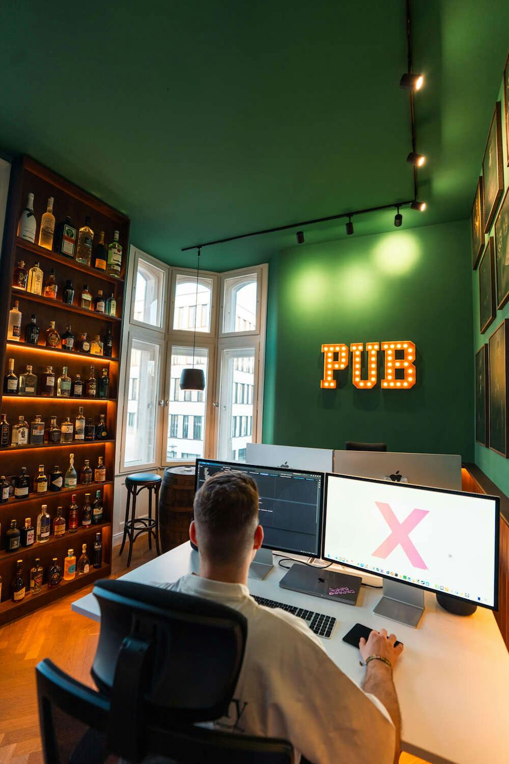 Arbeitsplatz im Exportarts Pub Büro mit Alkoholwand, grünen Wänden und beleuchtetem PUB Schriftzug Arbeitsplatz im Exportarts Pub Büro mit Alkoholwand, grünen Wänden und beleuchtetem PUB Schriftzug