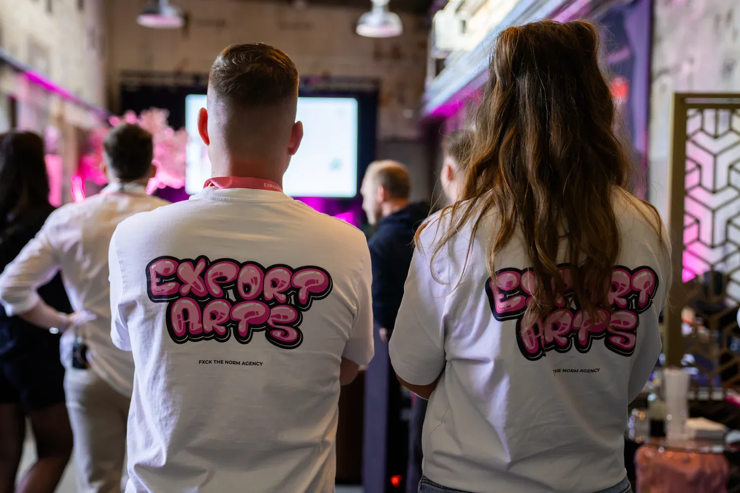 Zwei Personen von hinten in weißen Exportarts-Shirts mit pinkem Graffiti-Logo beim Vortrag
