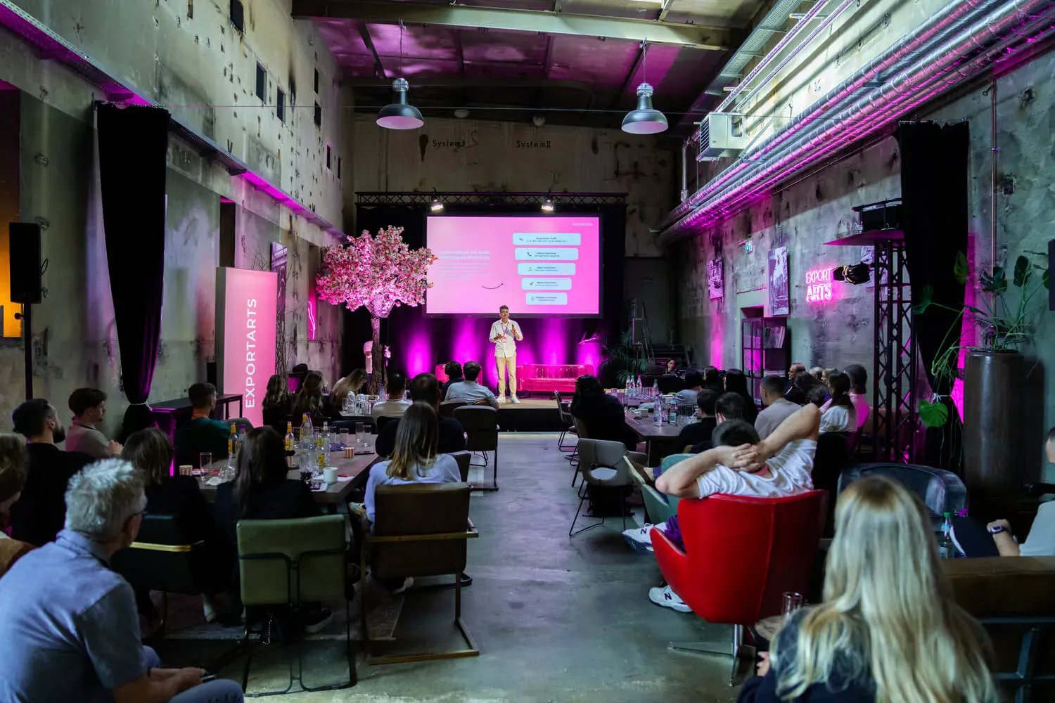 Publikum verfolgt Vortrag in Industrielocation mit pinker Bühne und Exportarts-Branding