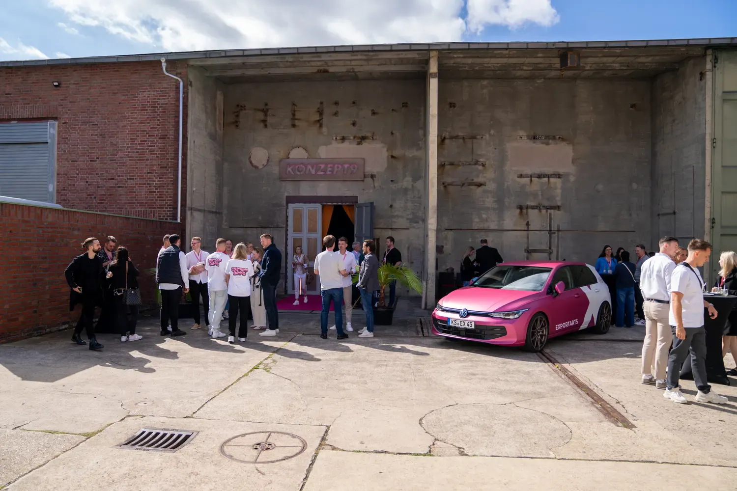 Eventgäste im Außenbereich vor Industriehalle mit pinkem VW Golf als Eyecatcher