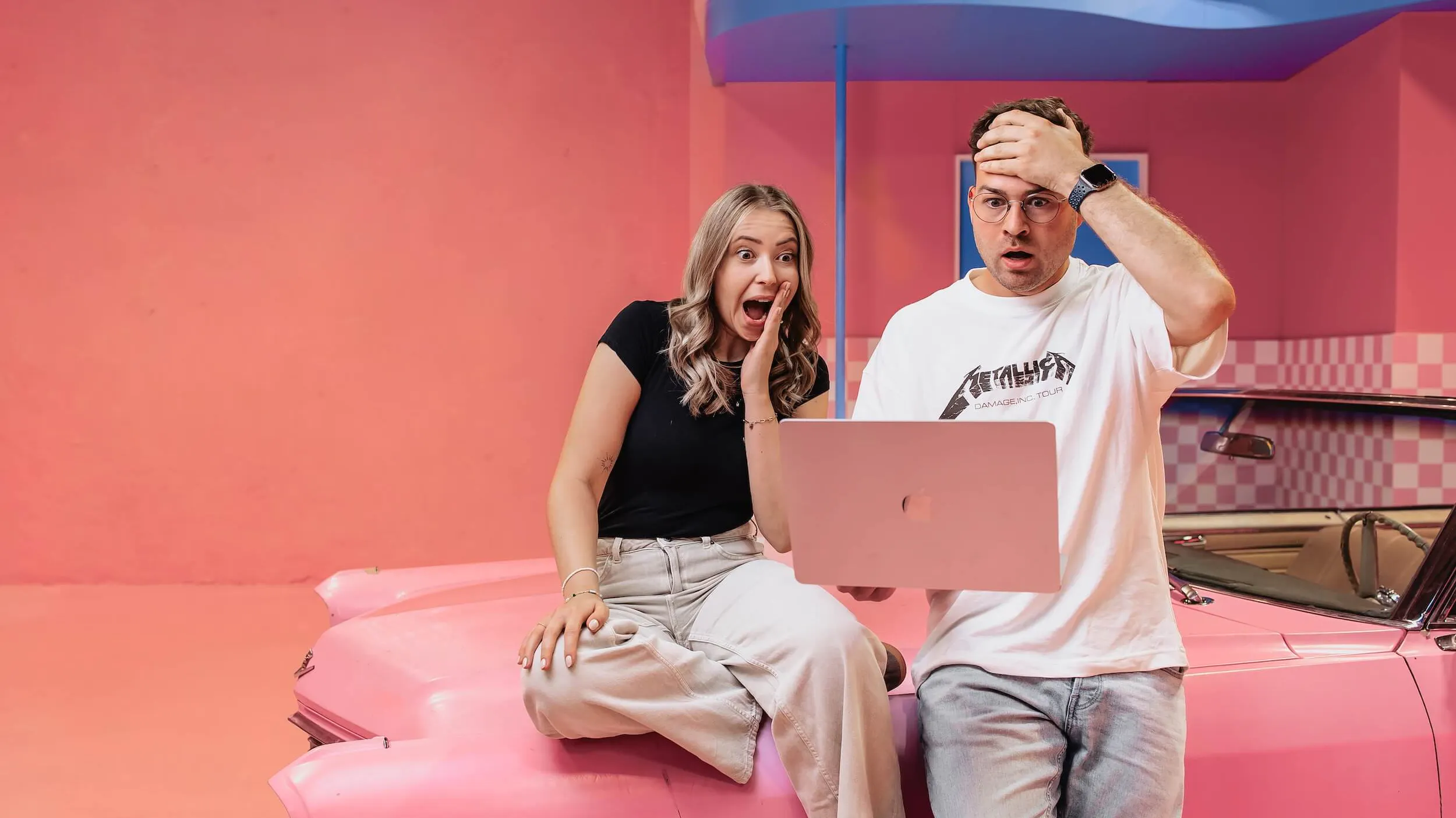 Zwei Mitarbeiter der SEO Copywriting Agentur sitzen auf einem pinken Auto und schauen schockiert in ein Macbook