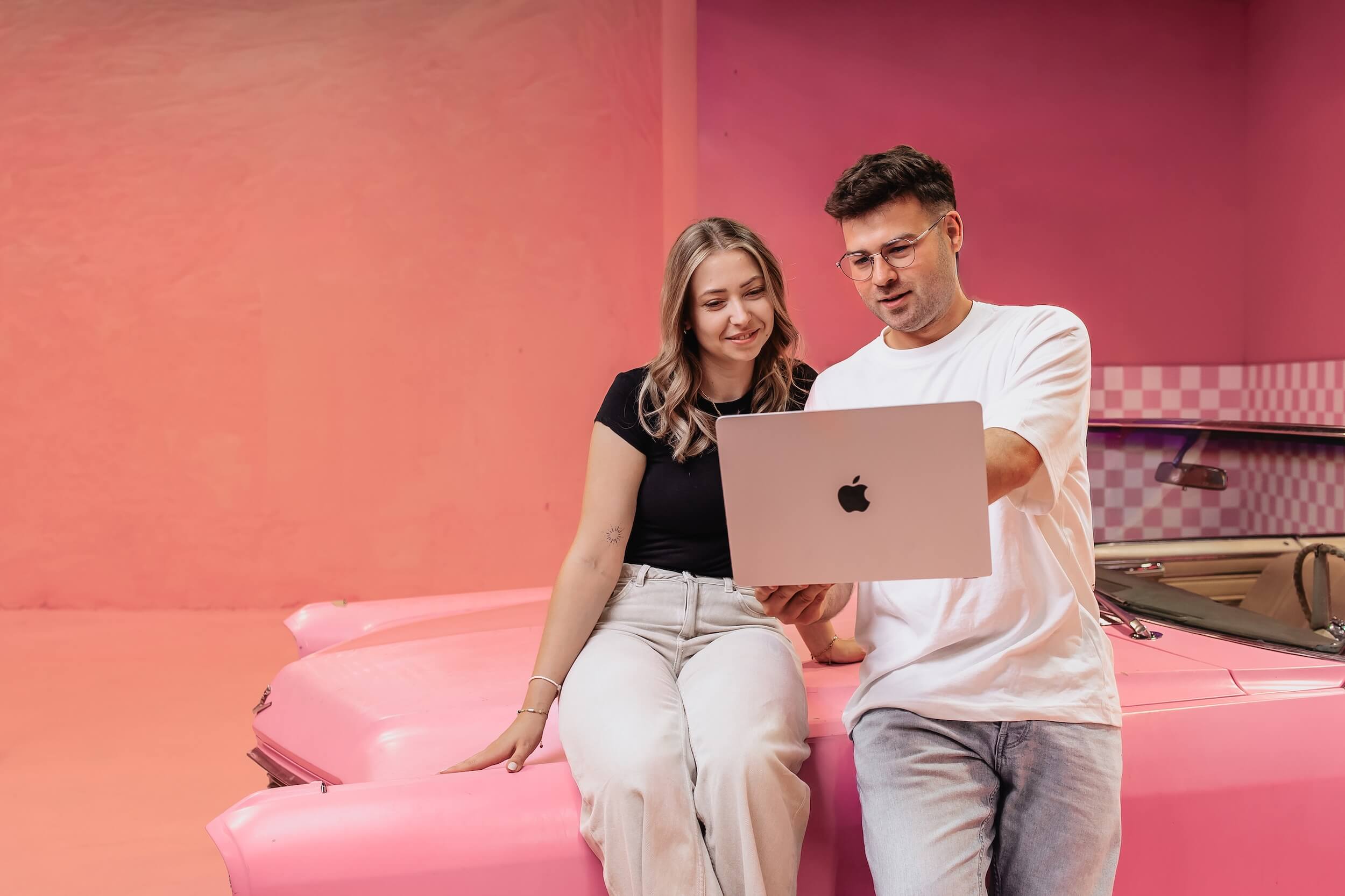 Zwei Mitarbeiter der Contentful Agentur Exportarts sitzen auf einem pinken Auto und schauen gemeinsam in ein Macbook