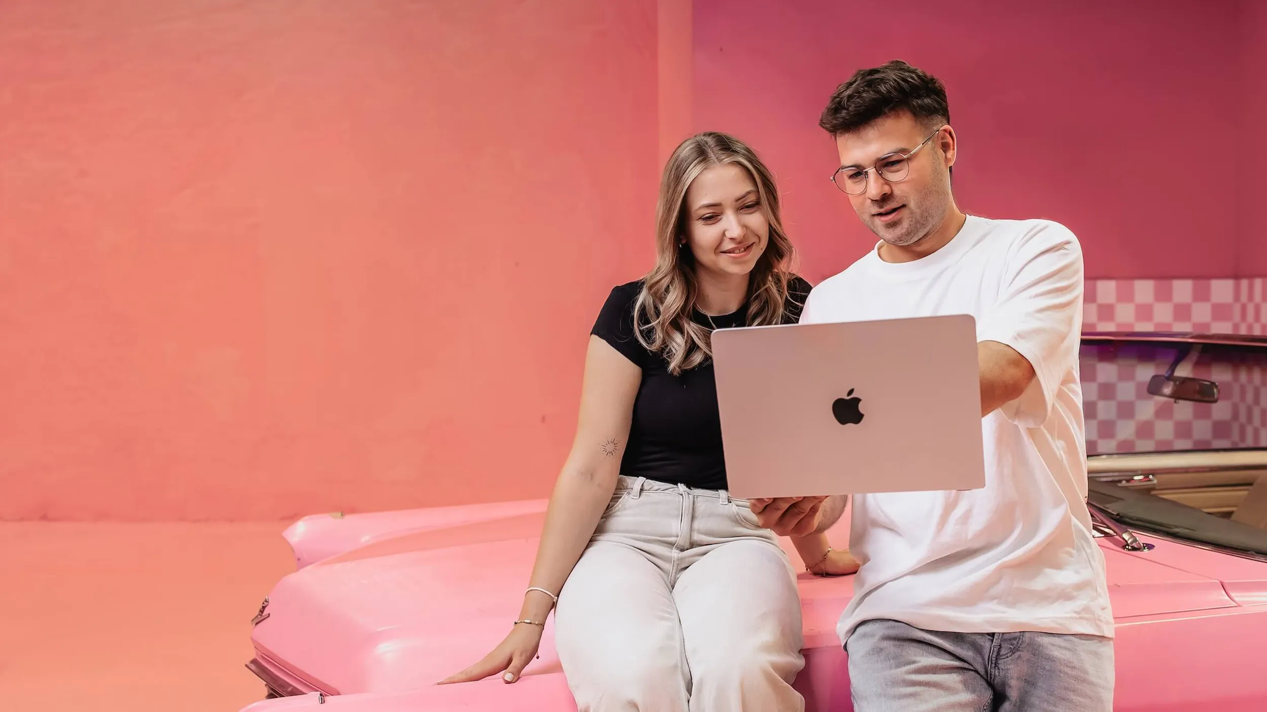 Zwei Mitarbeiter der Website Agentur Exportarts sitzen auf einem pinken Auto und schauen auf einen Laptop