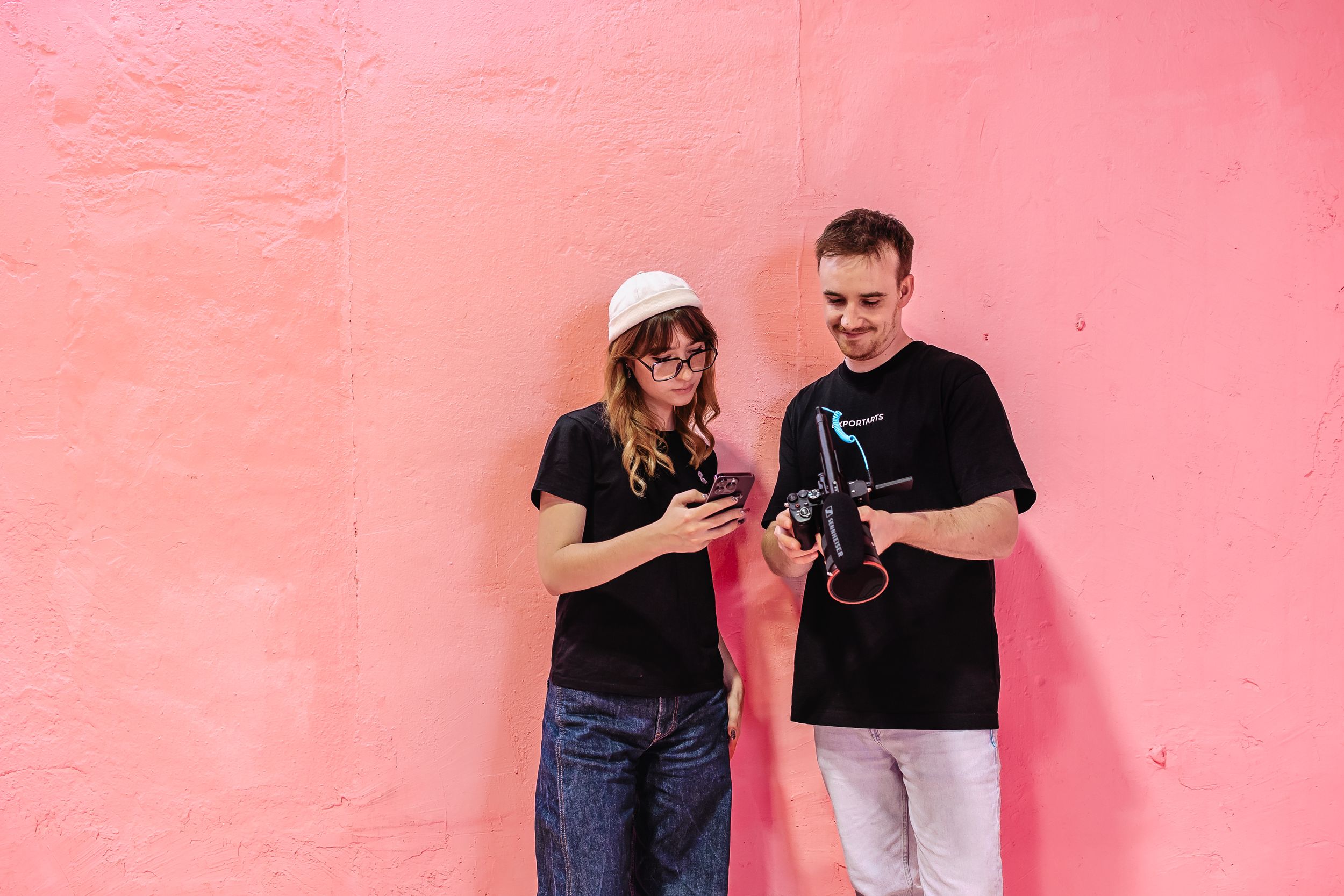 Zwei Mitarbeiter der Website Agentur Exportarts stehen vor einer pinken Wand und schauen in ein Handy und eine Kamera