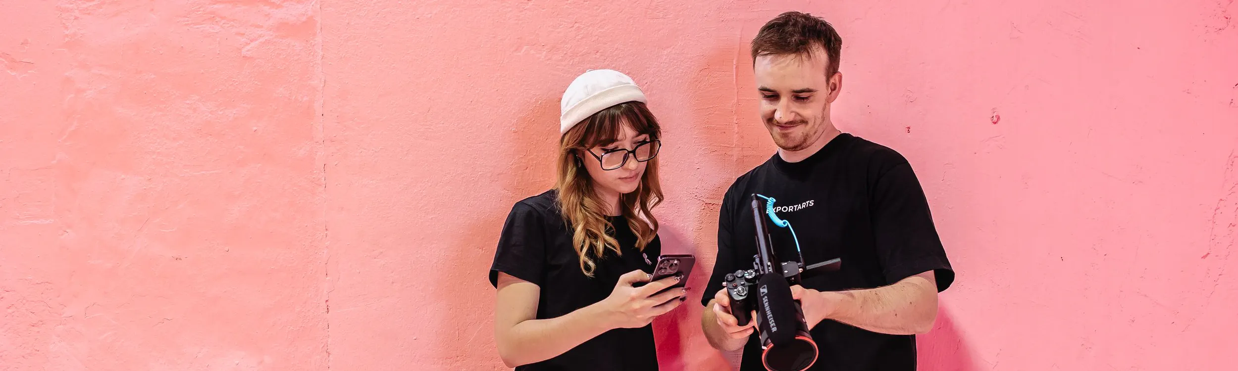 Zwei Mitarbeiter der Content Agentur Exportarts mit iPhone und Kamera vor einer pinken Wand