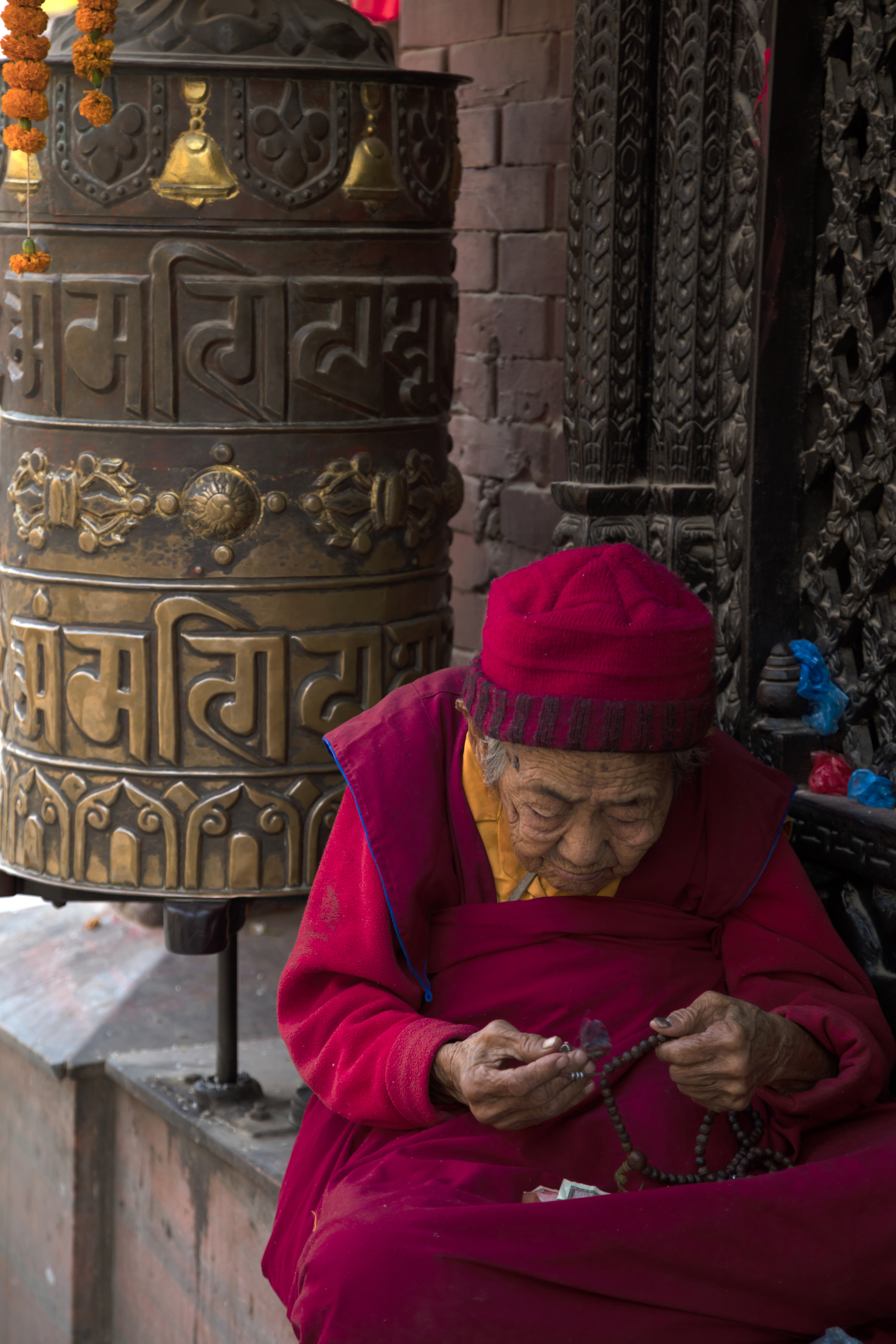 Elderly devotee praying with beads beside a prayer wheel in Kathmandu, Nepal
