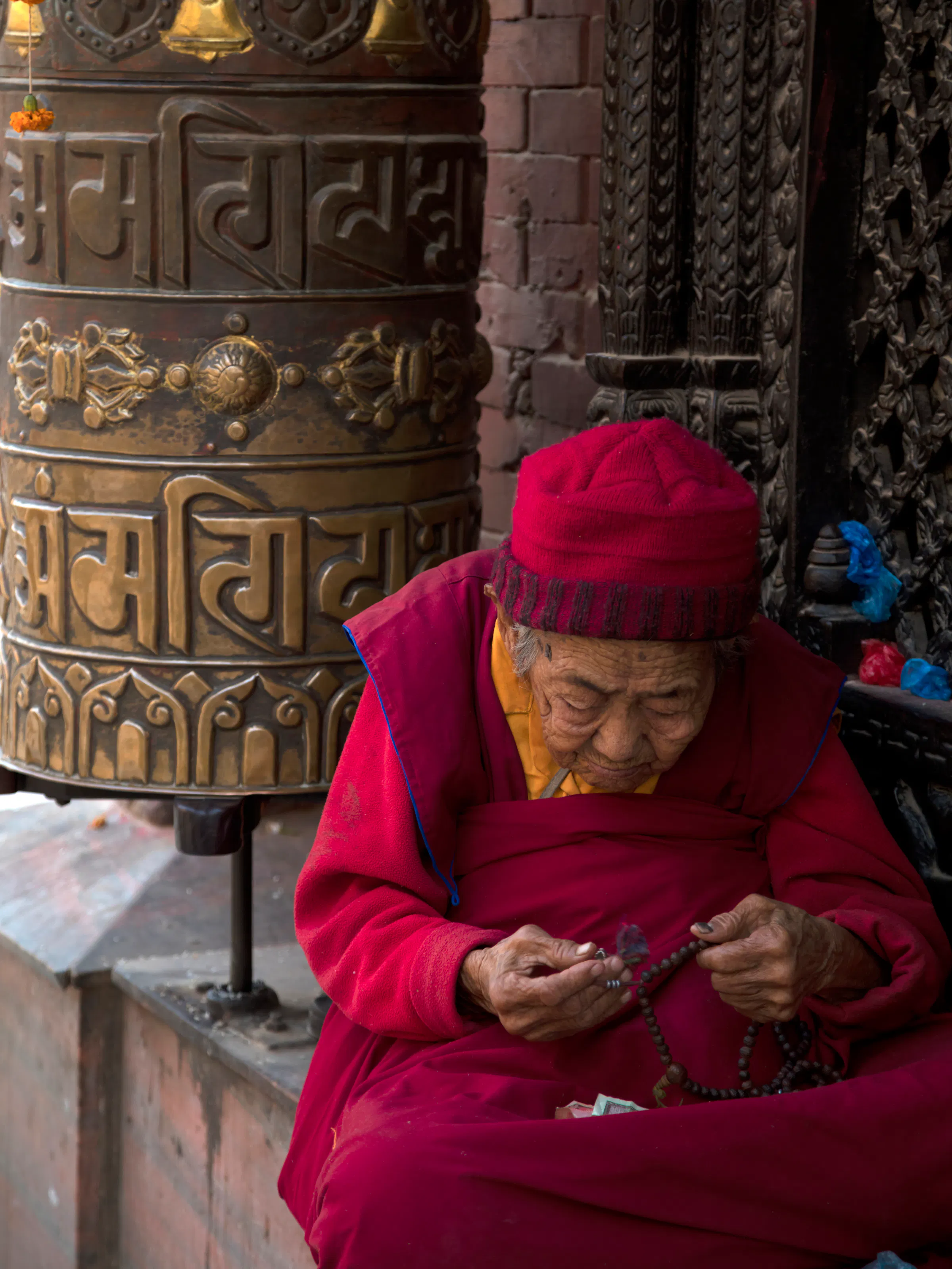 Elderly devotee praying with beads beside a prayer wheel in Kathmandu, Nepal