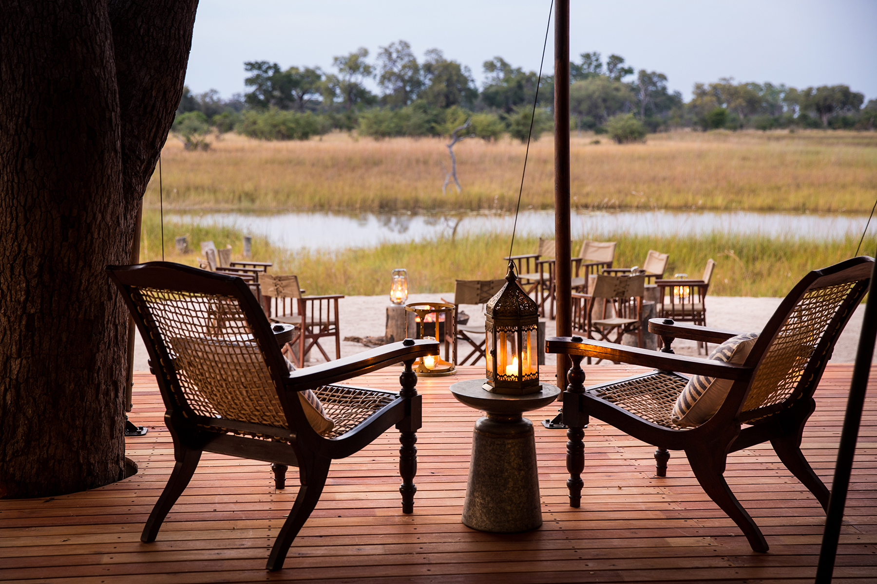 Wicker armchairs and lantern light on a wooden deck overlooking the Okavango Delta floodplain at dusk, Botswana.