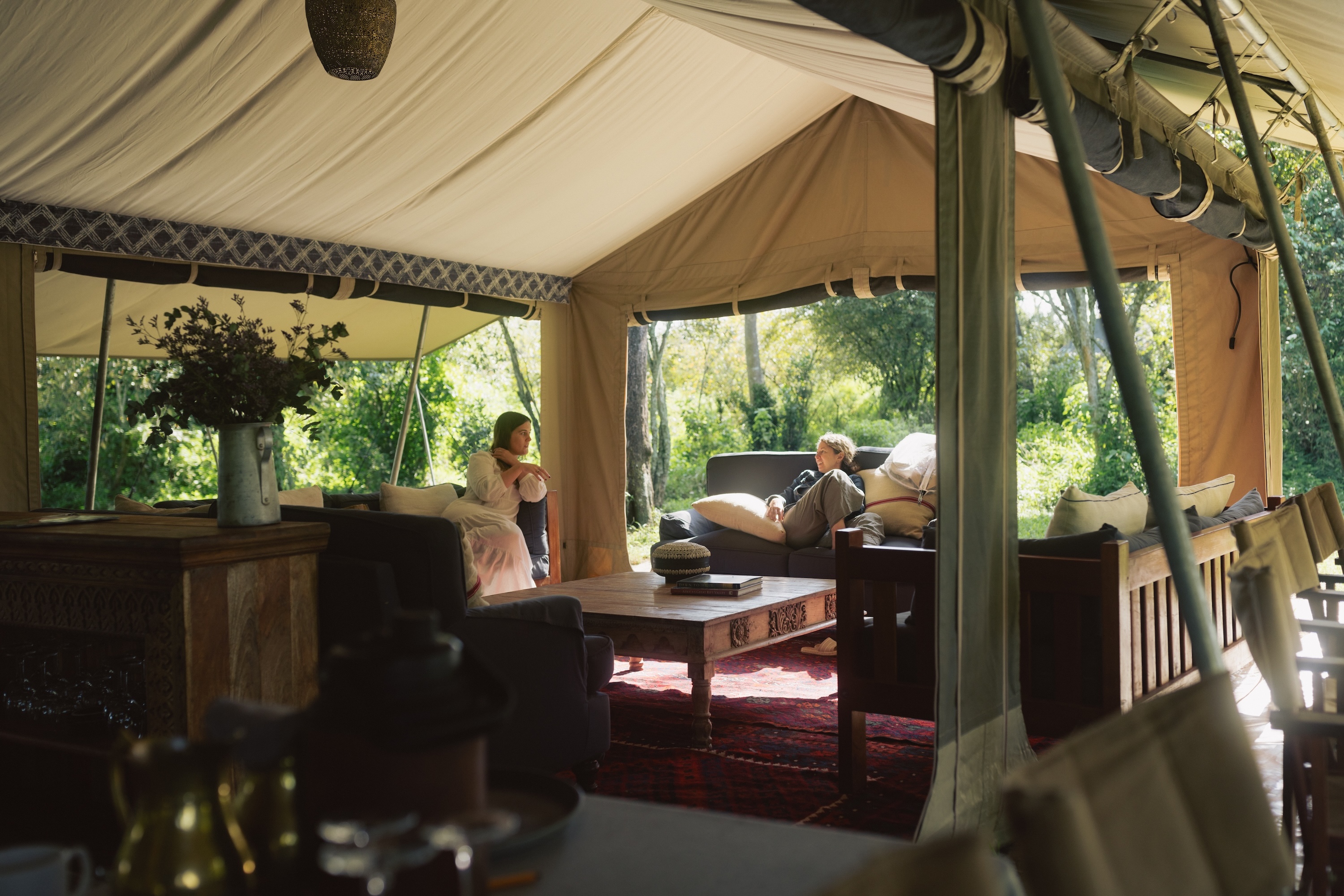 Two women chat on outdoor sofas in a safari lounge tent surrounded by lush greenery.