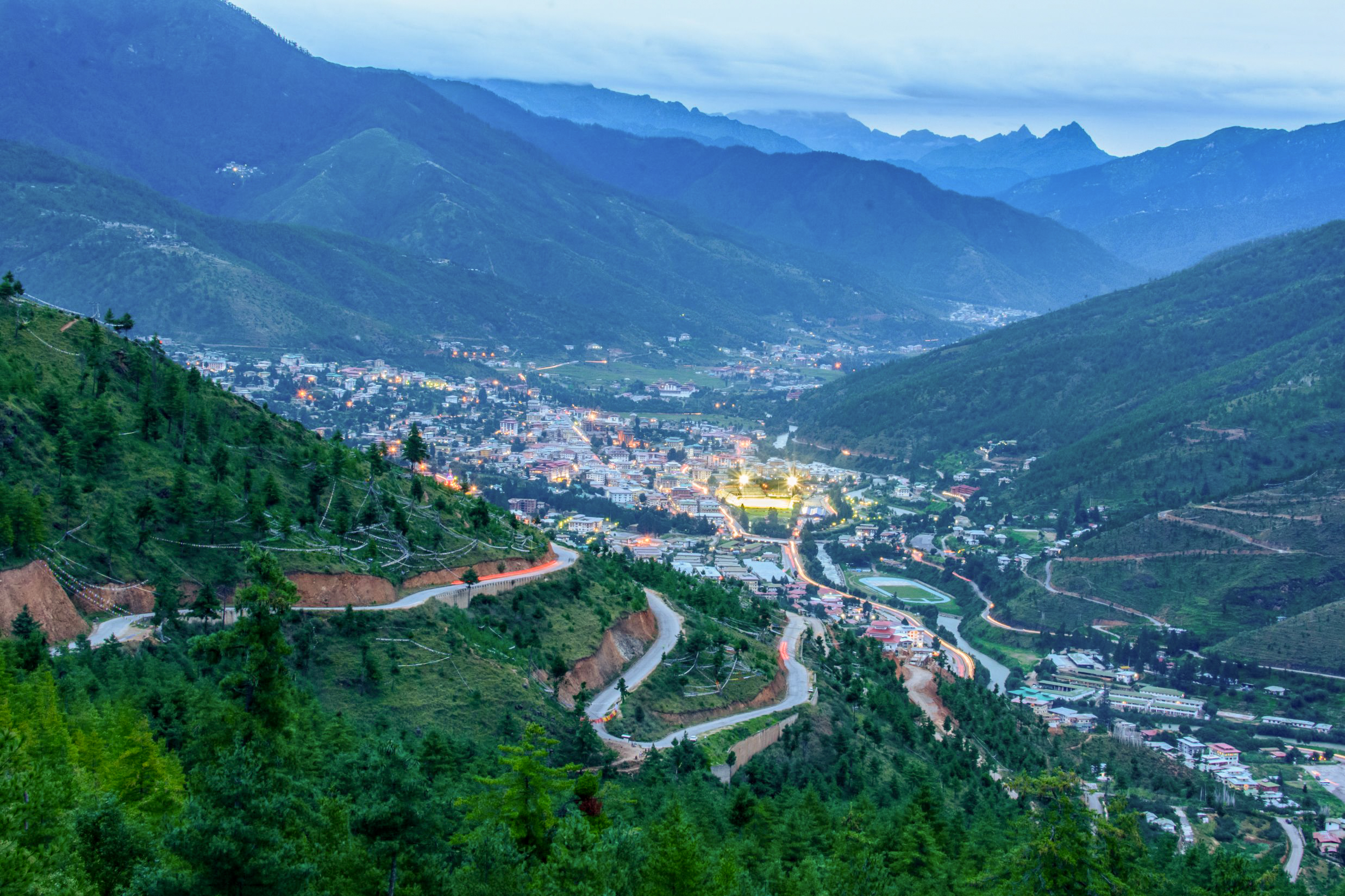 Aerial twilight view of Thimphu, Bhutan, nestled in a forested Himalayan valley. City lights illuminate the valley floor, and switchback roads cut down the pine-covered mountainside in the foreground, with layered blue mountain ridges fading into the misty distance.