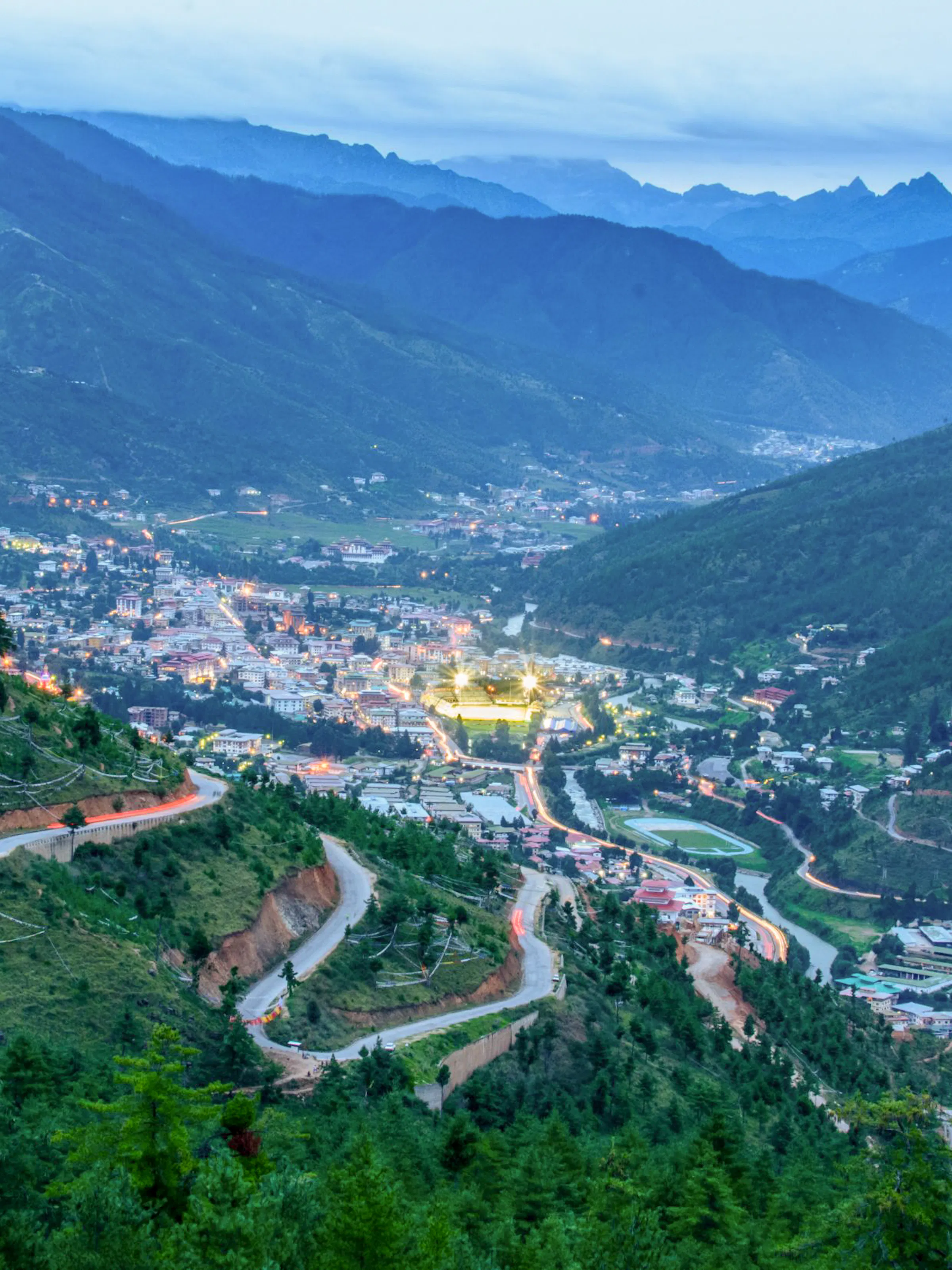 Aerial twilight view of Thimphu, Bhutan, nestled in a forested Himalayan valley. City lights illuminate the valley floor, and switchback roads cut down the pine-covered mountainside in the foreground, with layered blue mountain ridges fading into the misty distance.
