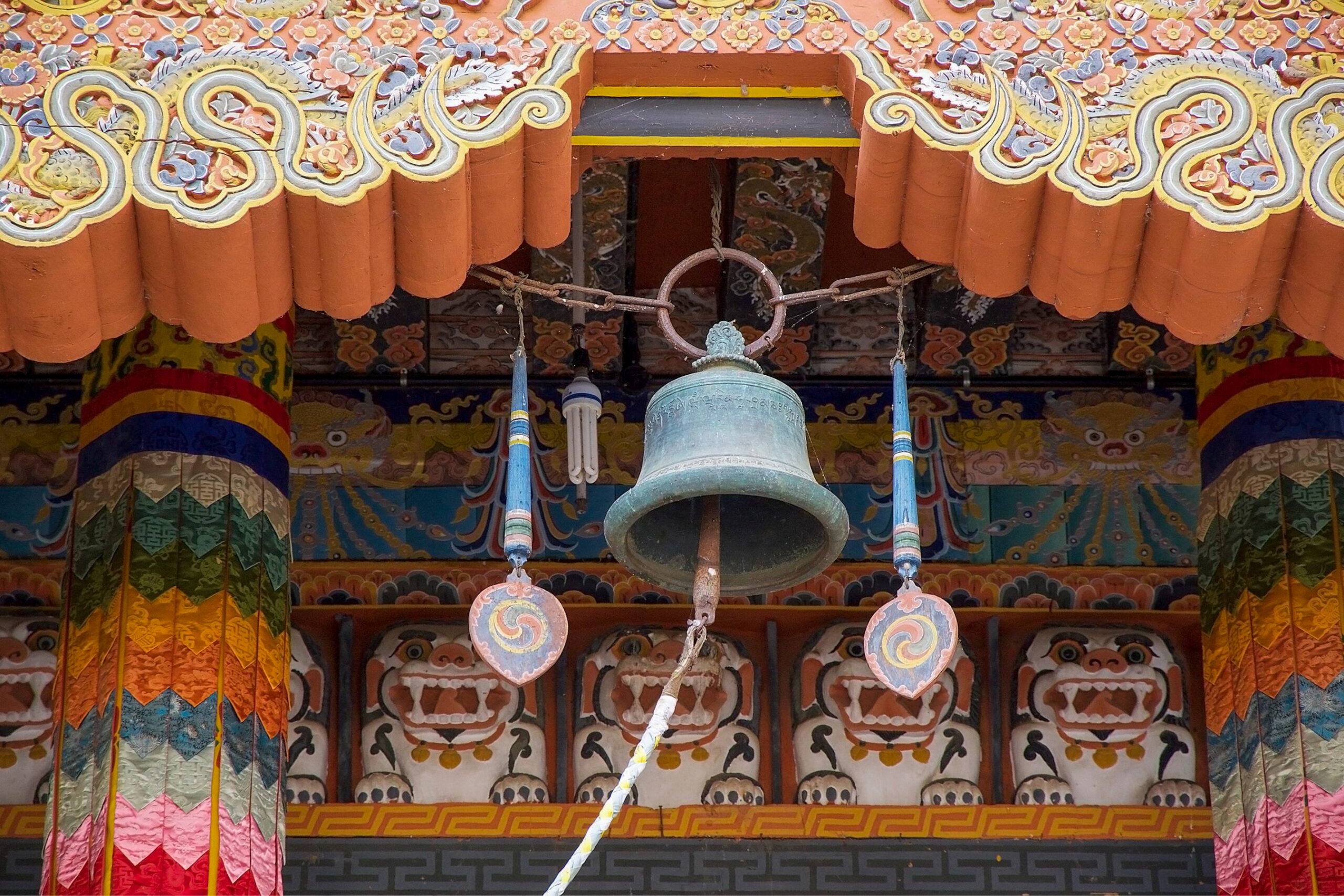 A large bronze temple bell hangs from an ornately carved wooden ceiling at Punakha Dzong in Bhutan, framed by hand-painted lotus, cloud, and dragon motifs in orange, gold, and blue.