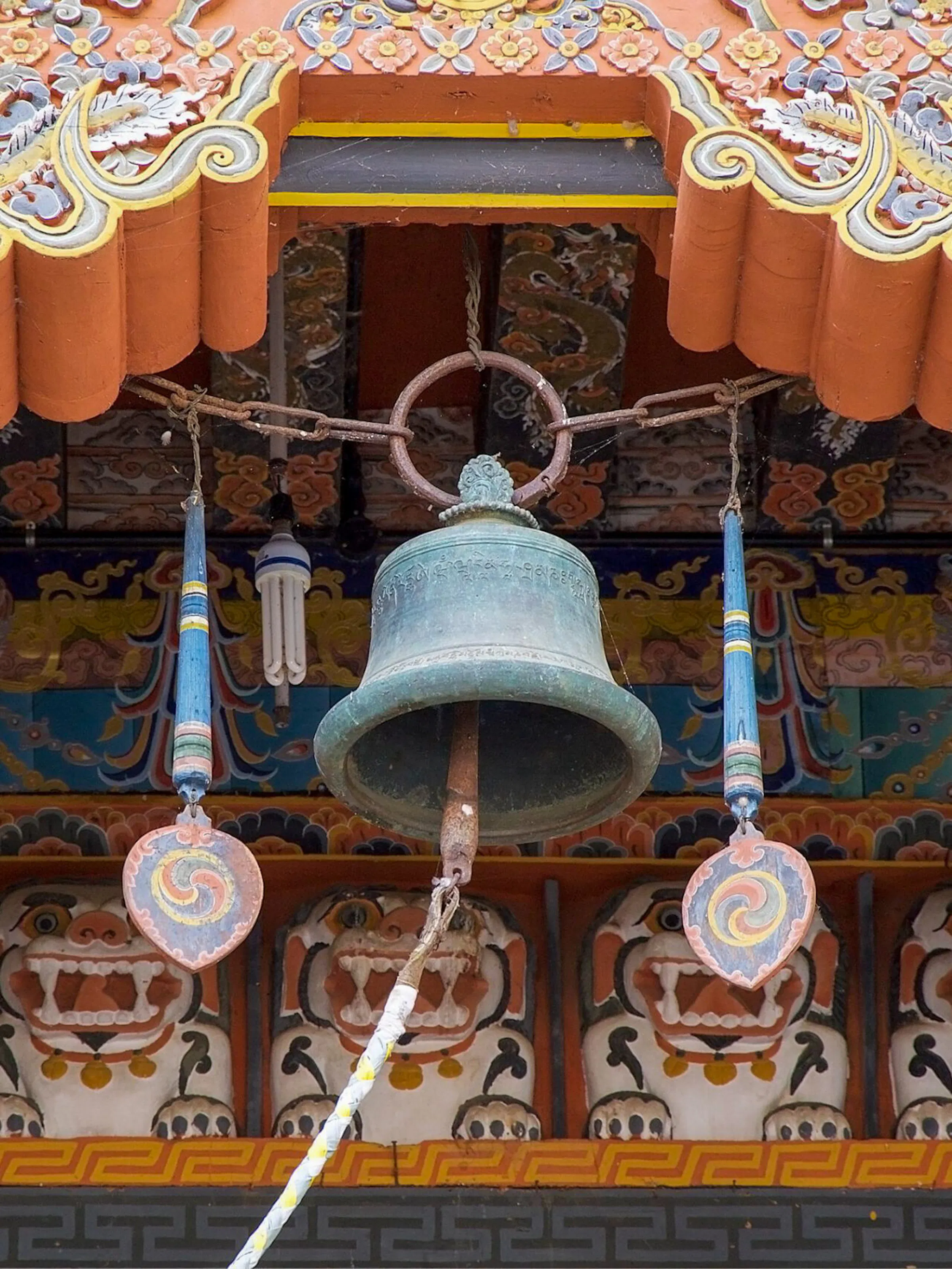 A large bronze temple bell hangs from an ornately carved wooden ceiling at Punakha Dzong in Bhutan, framed by hand-painted lotus, cloud, and dragon motifs in orange, gold, and blue.