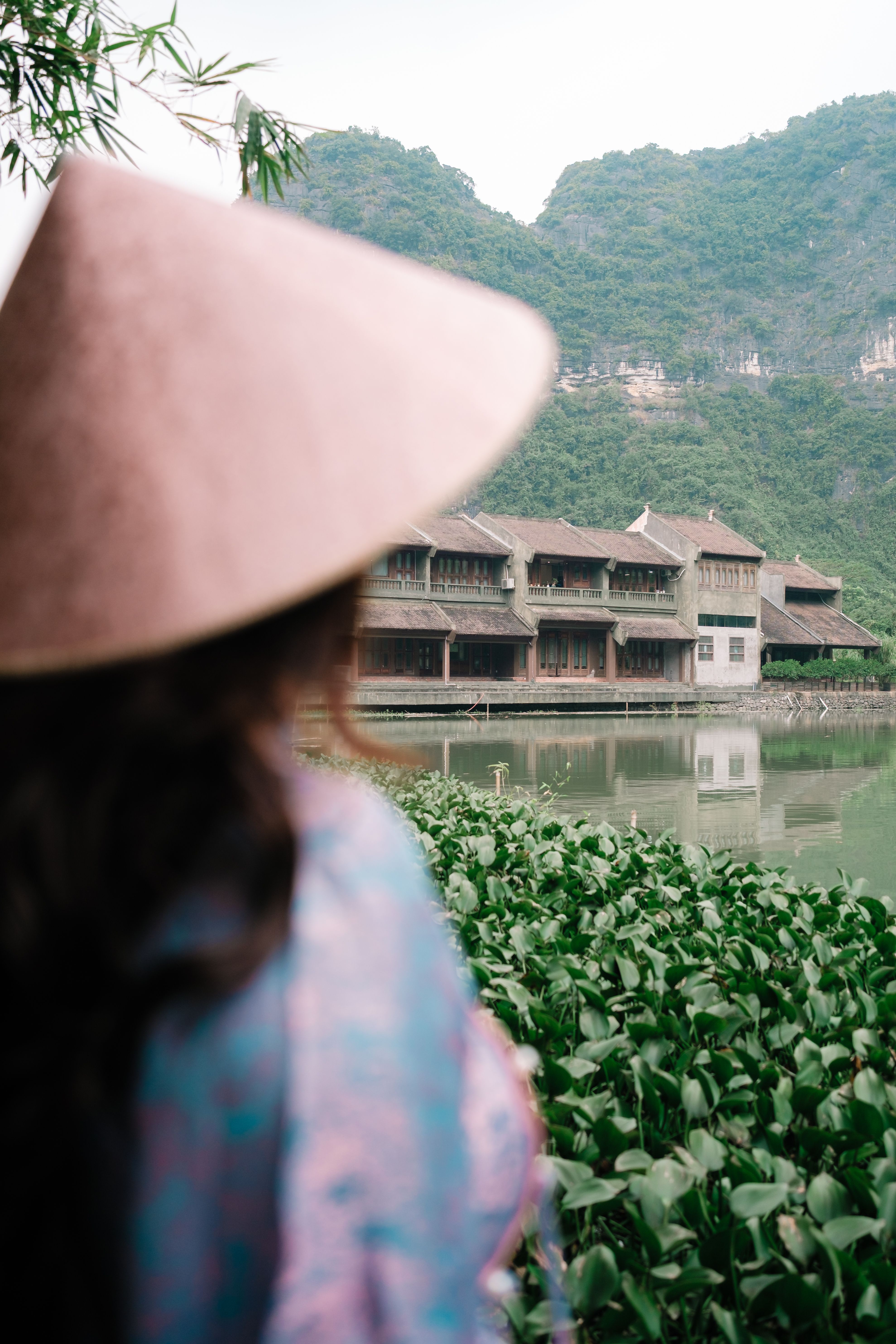 Woman in conical hat beside traditional waterfront resort, Ninh Binh Vietnam