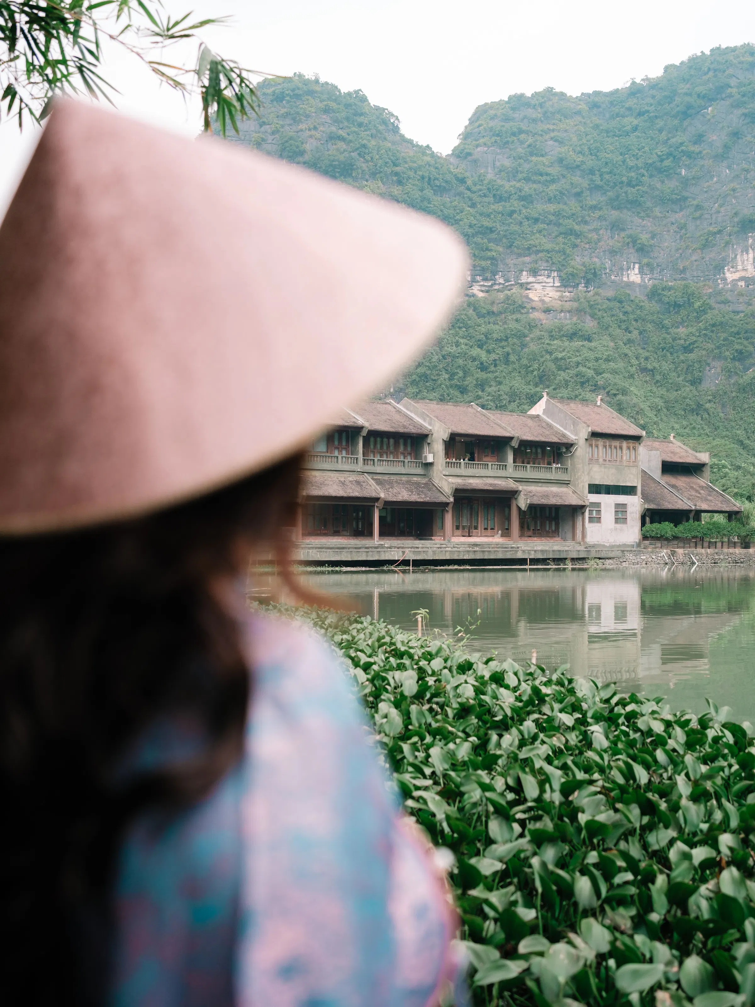 Woman in conical hat beside traditional waterfront resort, Ninh Binh Vietnam