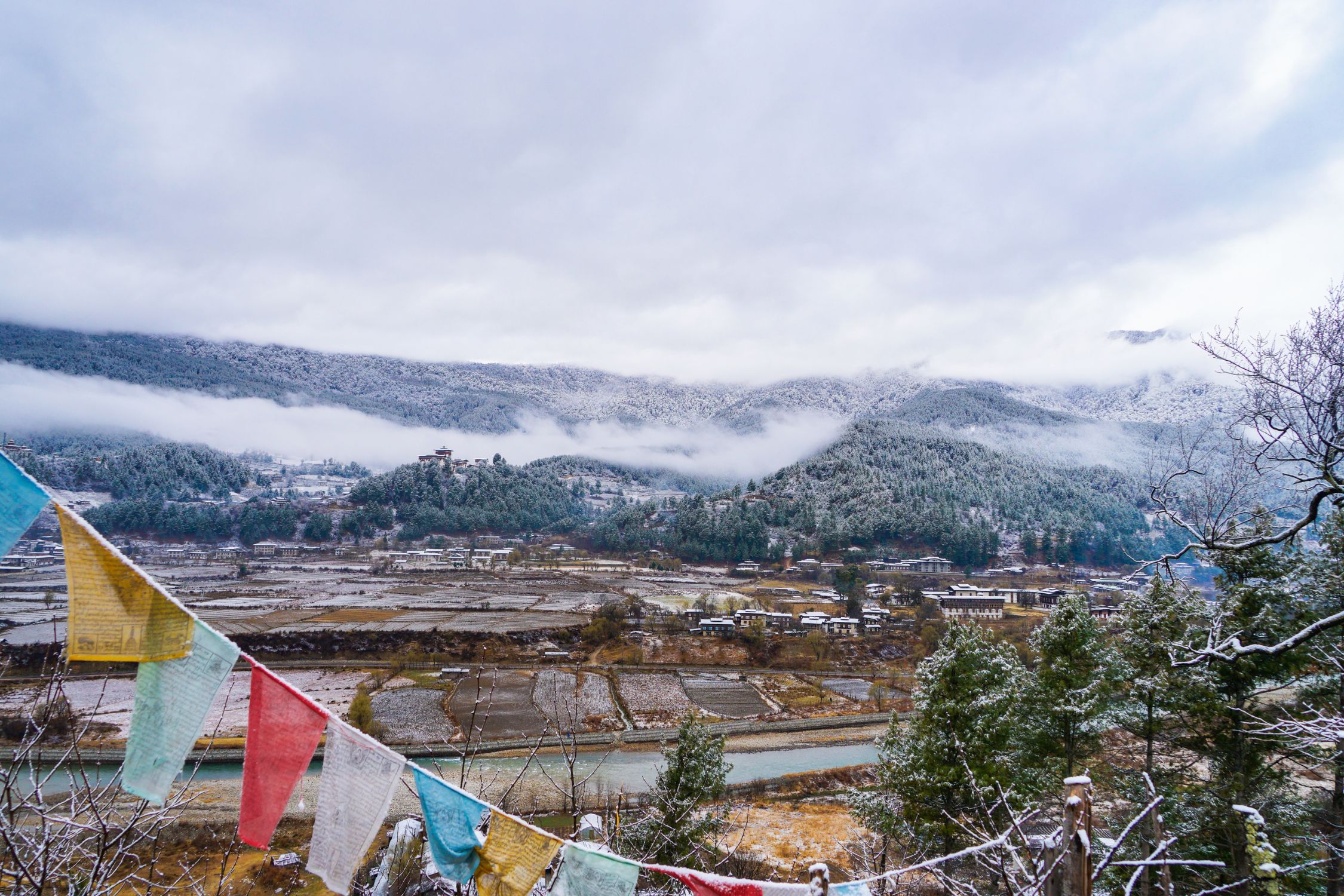 Prayer flags in blue, yellow, white, green, and red hang in the foreground, framing a wide valley view in Bhutan. Below, a river winds past patchwork fields dusted with snow.