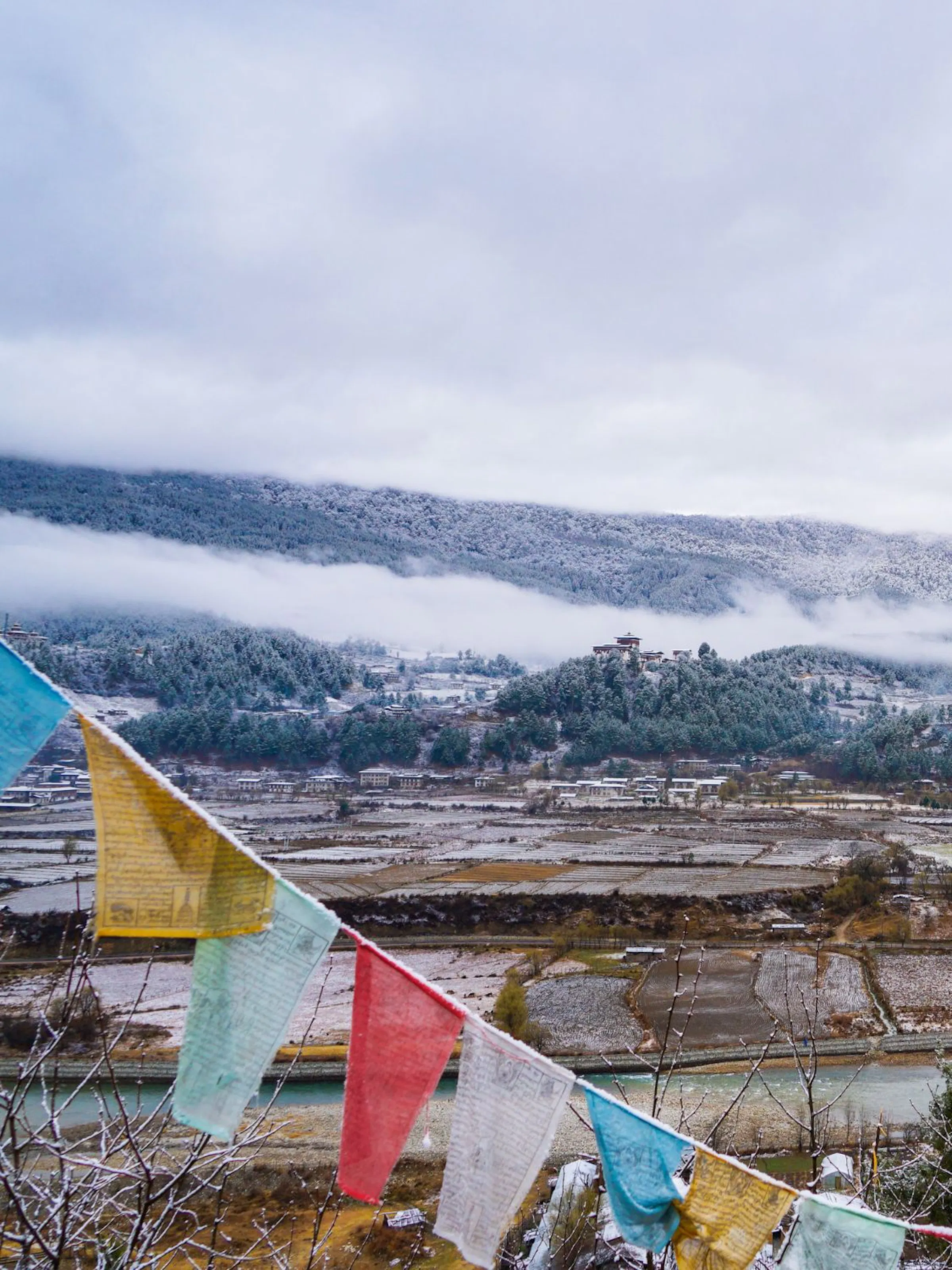 Prayer flags in blue, yellow, white, green, and red hang in the foreground, framing a wide valley view in Bhutan. Below, a river winds past patchwork fields dusted with snow.