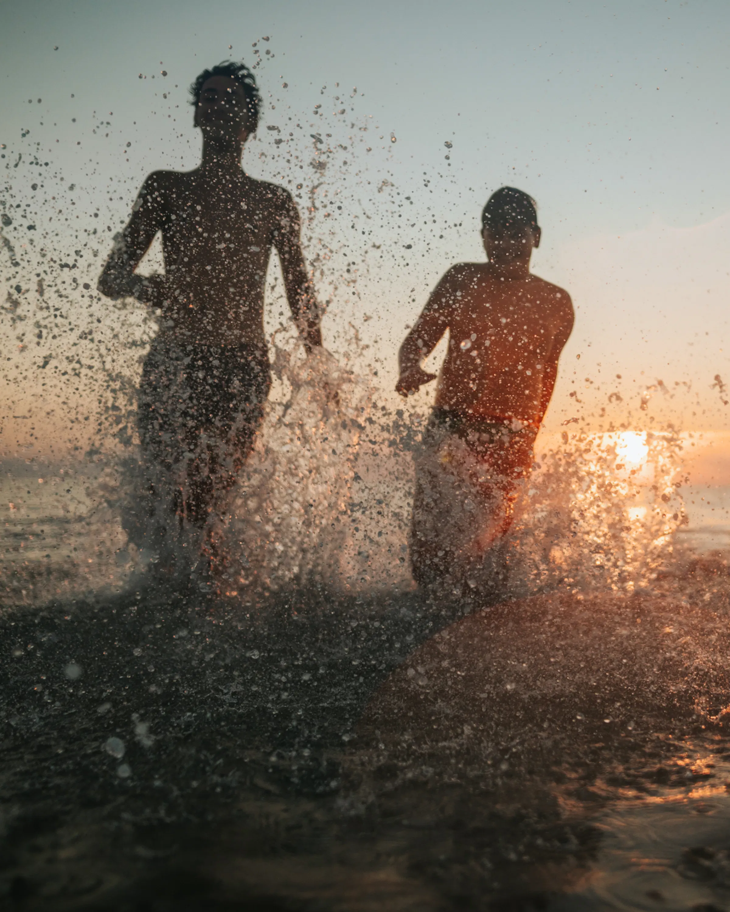 Two children run through ocean waves at sunset, with water splashing around them