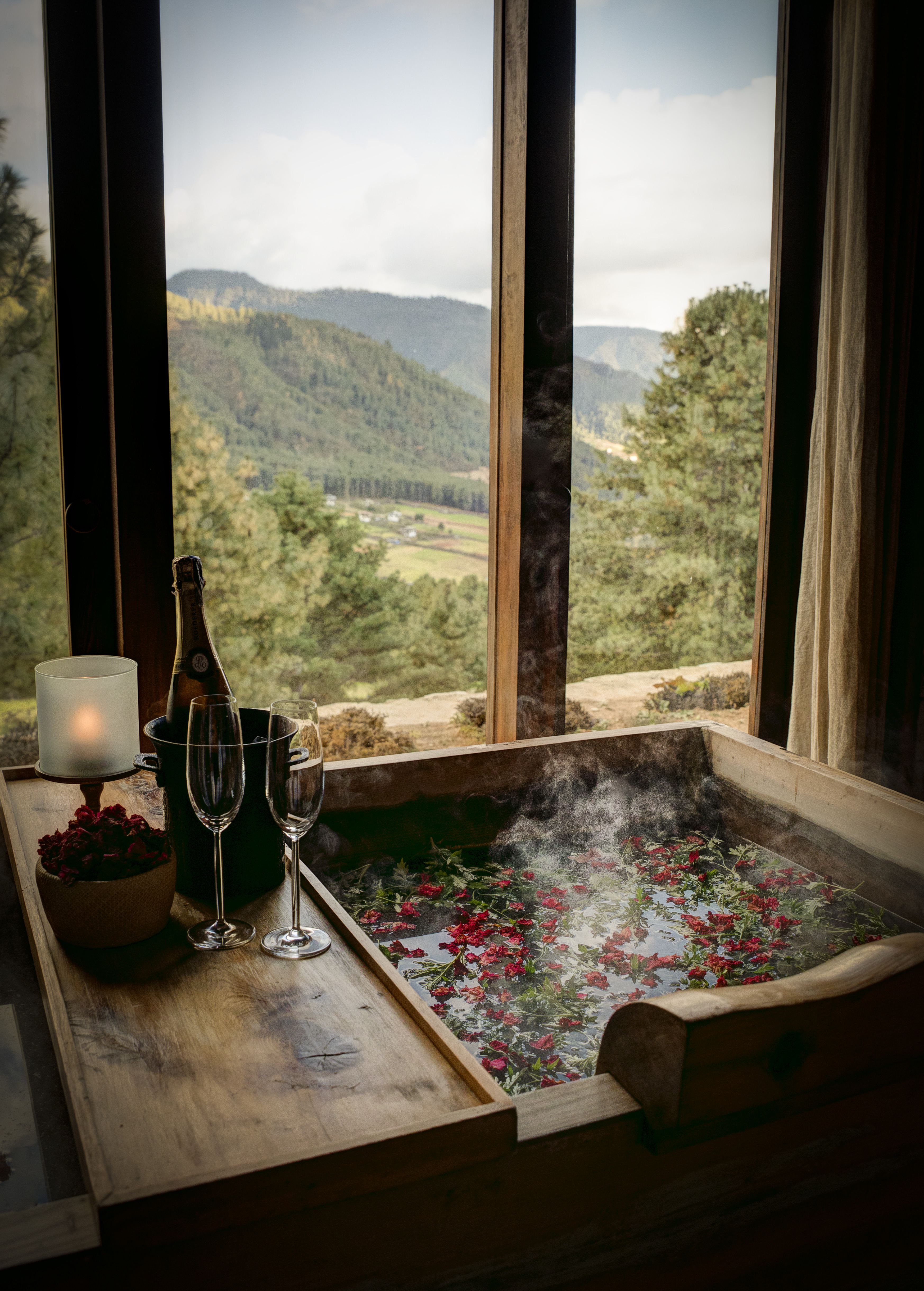 A wooden hot stone bath scattered with fresh flowers steams beside a window in Bhutan, overlooking pine-covered hills and a valley below.