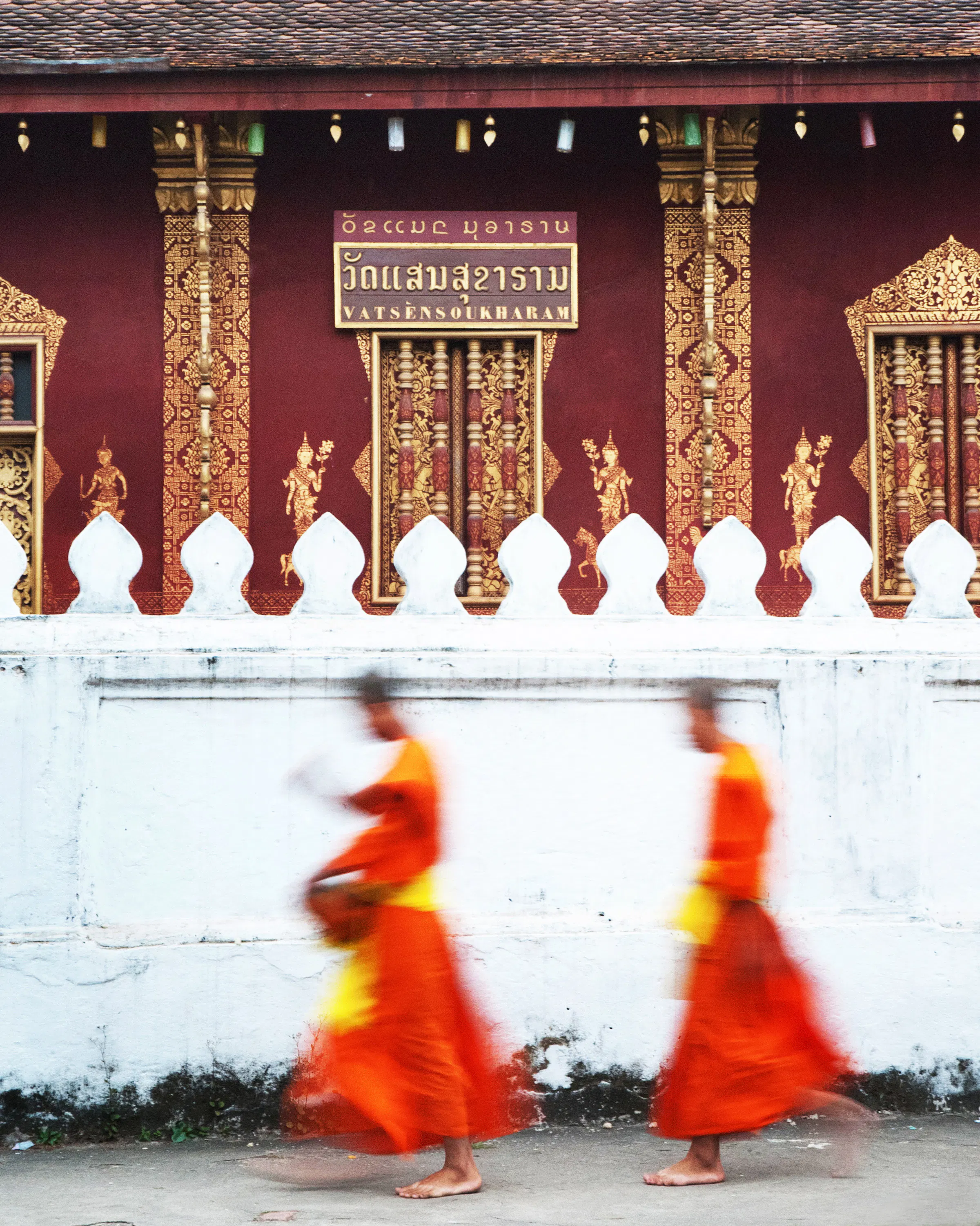 Two monks in orange robes walk past a temple in Laos