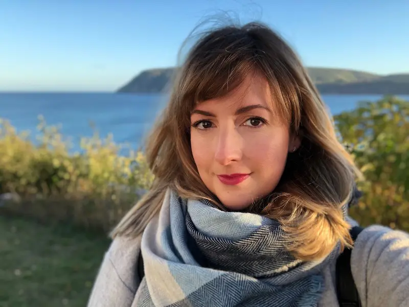 Portrait of Chloe Berge, writer at Extraordinary Journeys, standing outdoors near the coast, wearing a scarf with the ocean and cliffs in the background.