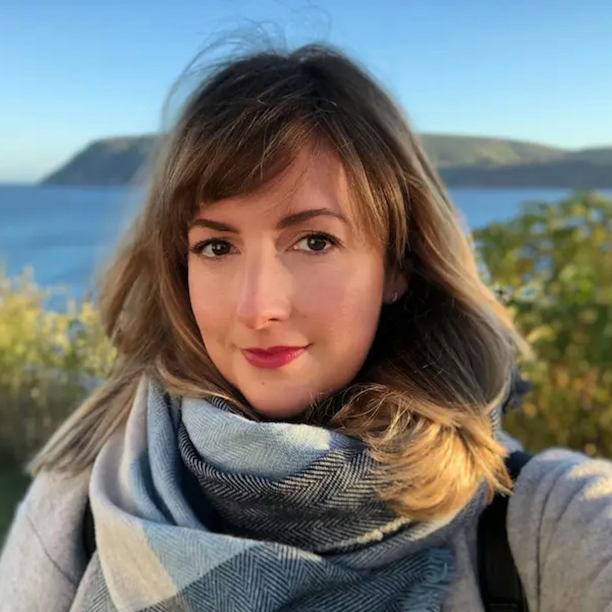 Portrait of Chloe Berge, writer at Extraordinary Journeys, standing outdoors near the coast, wearing a scarf with the ocean and cliffs in the background.