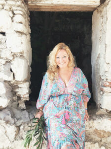 Vickie Paget, writer for Extraordinary Journeys, stands in a rustic stone doorway, wearing a colorful patterned dress and holding greenery.