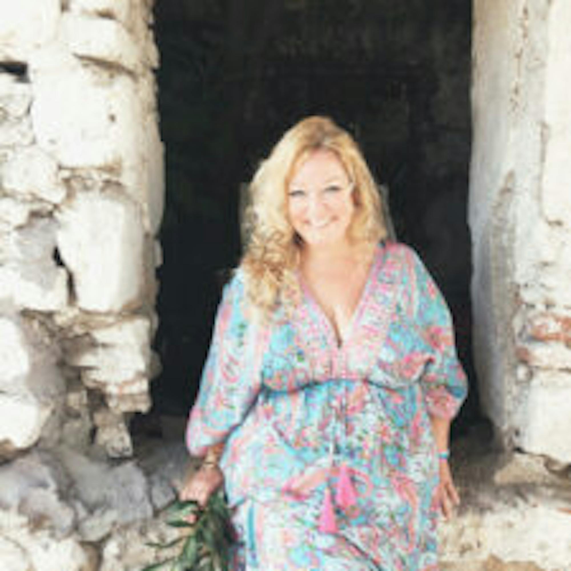 Vickie Paget, writer for Extraordinary Journeys, stands in a rustic stone doorway, wearing a colorful patterned dress and holding greenery.
