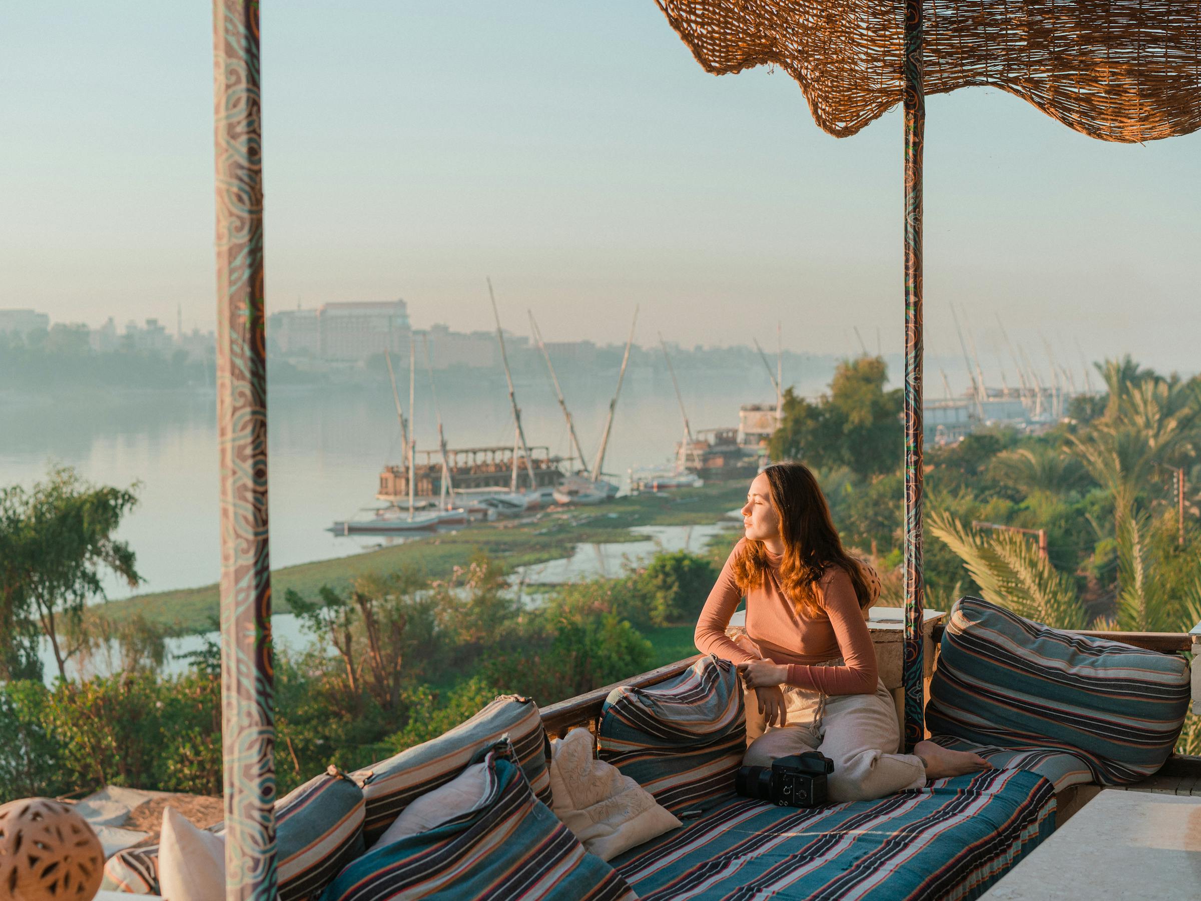 Person sits on patterned cushions under shade, overlooking a river with sailboats in the distance.