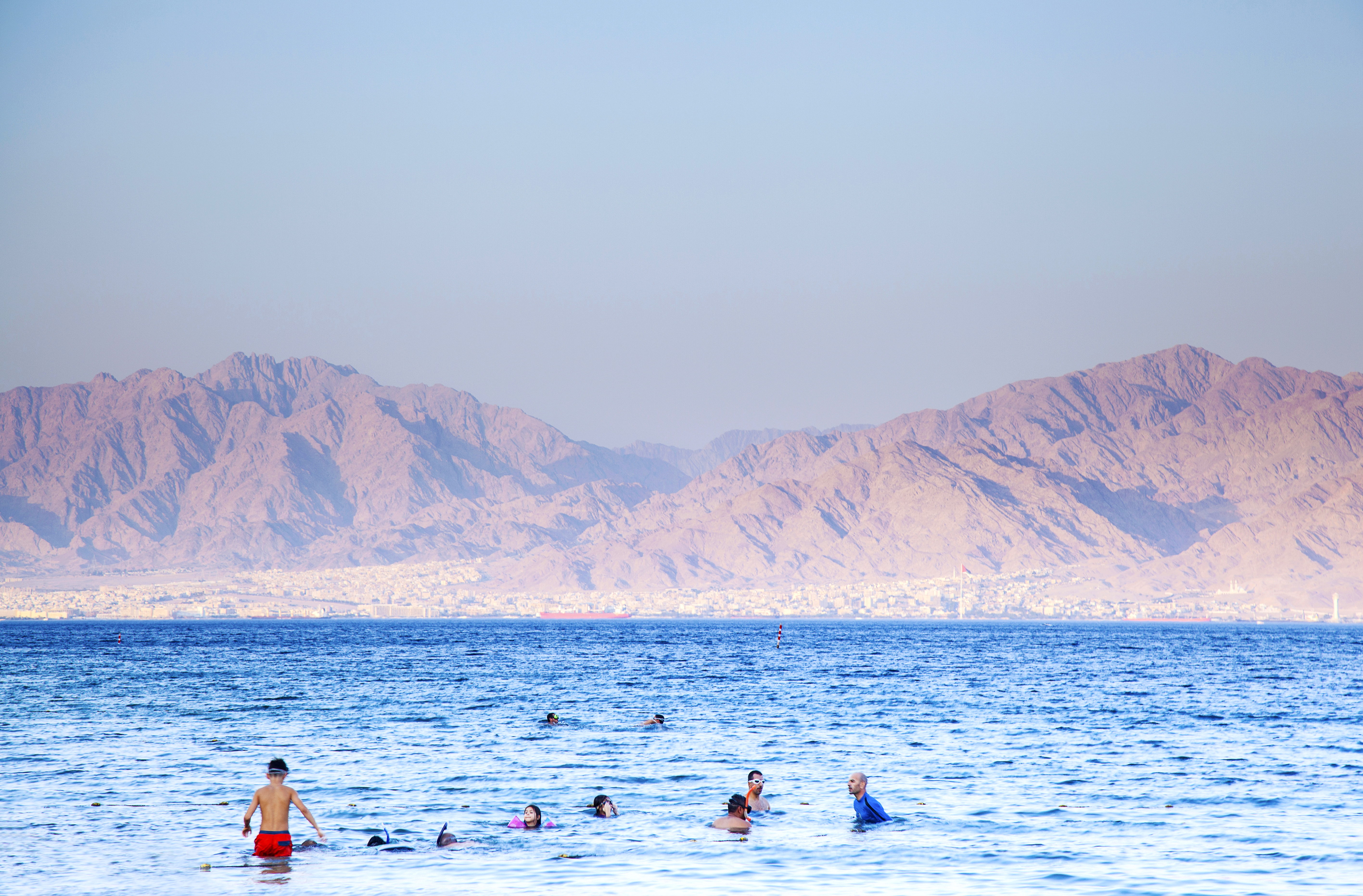 People swimming in clear blue water with rugged mountains on the horizon.