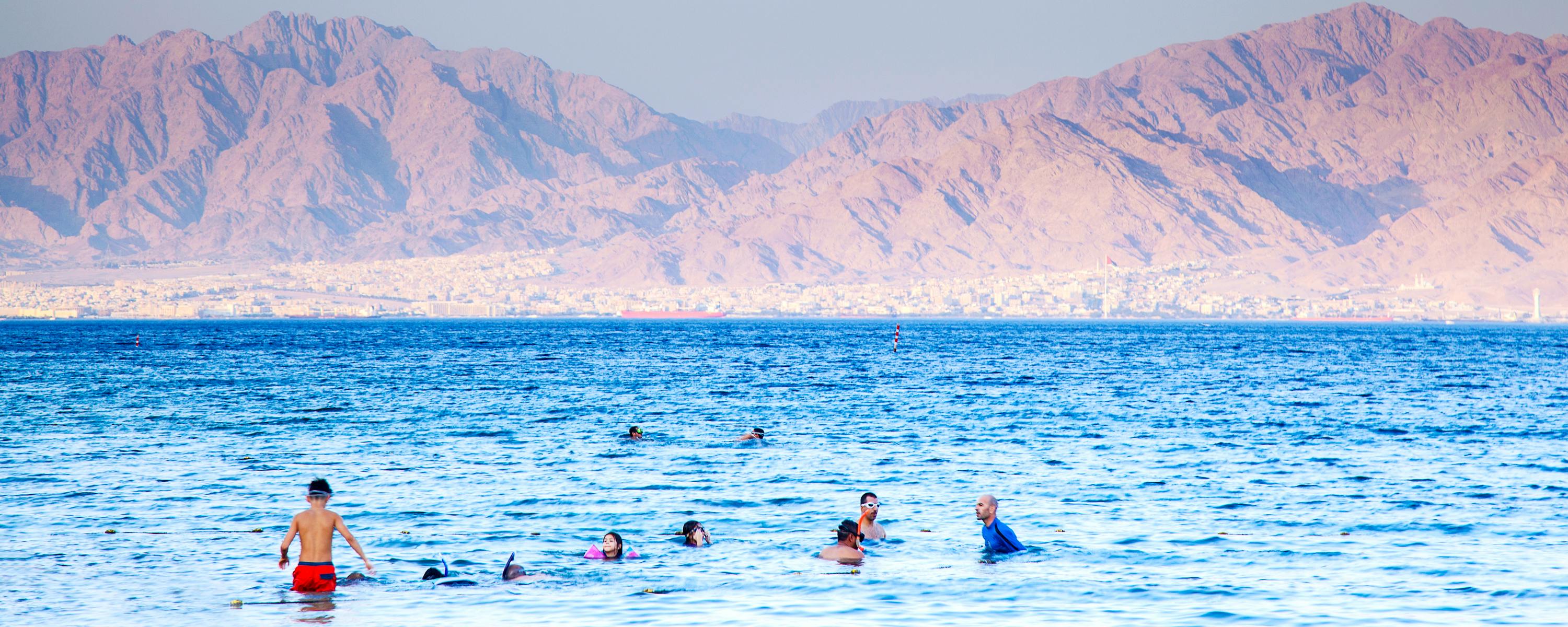 People swimming in clear blue water with rugged mountains on the horizon.