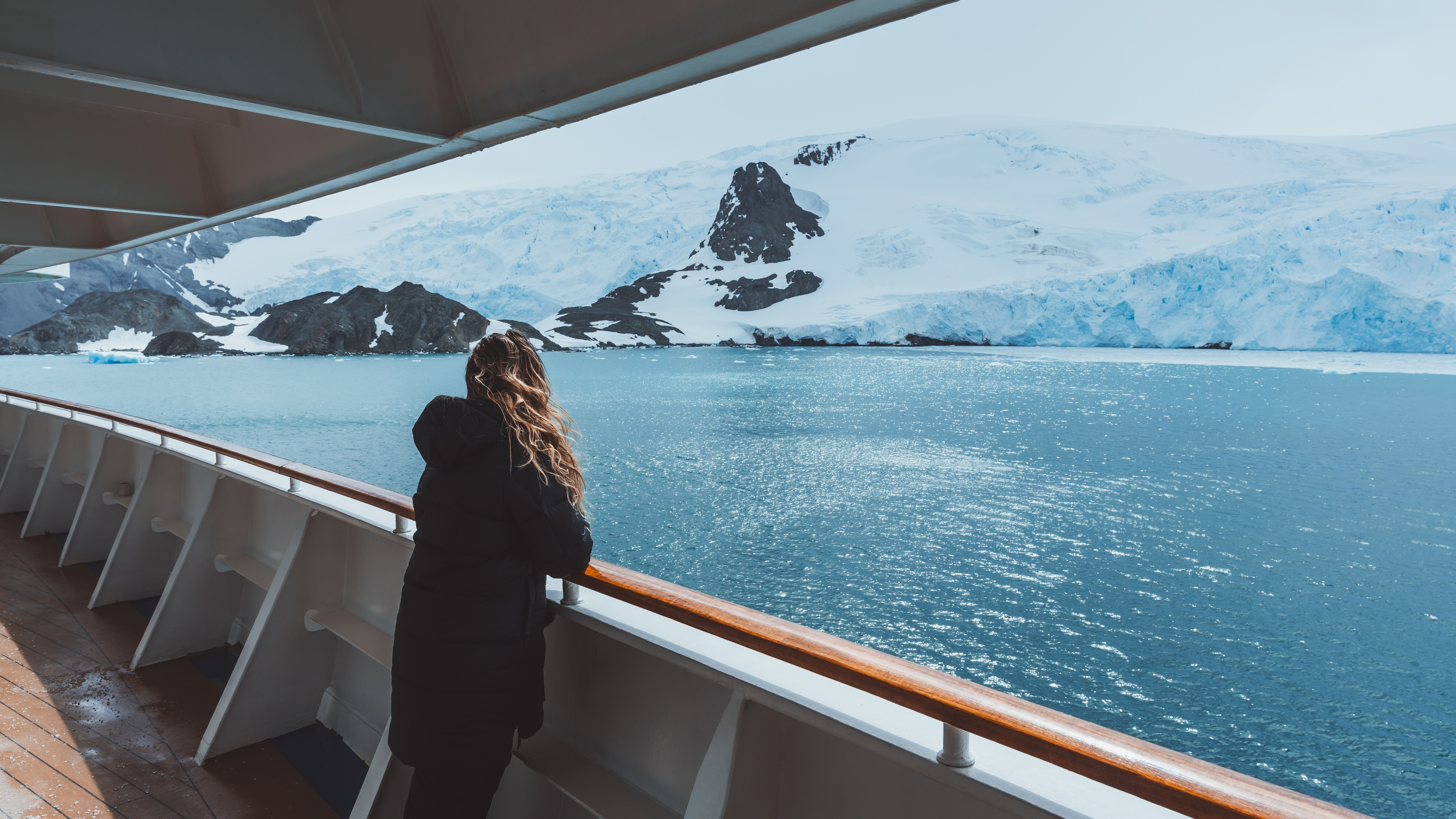 A female cruise passenger leans on a ship railing, watching glaciers and icy peaks across the Antarctic sea.