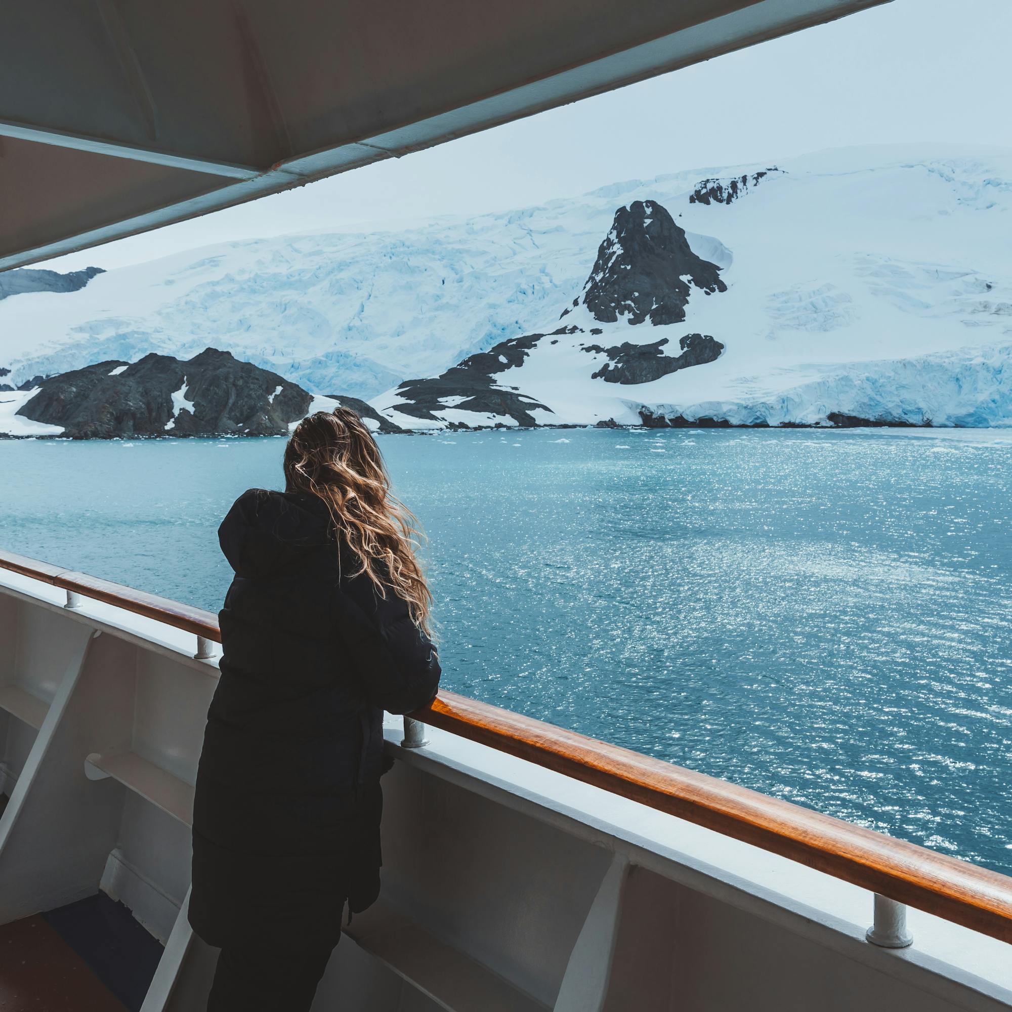 A female cruise passenger leans on a ship railing, watching glaciers and icy peaks across the Antarctic sea.