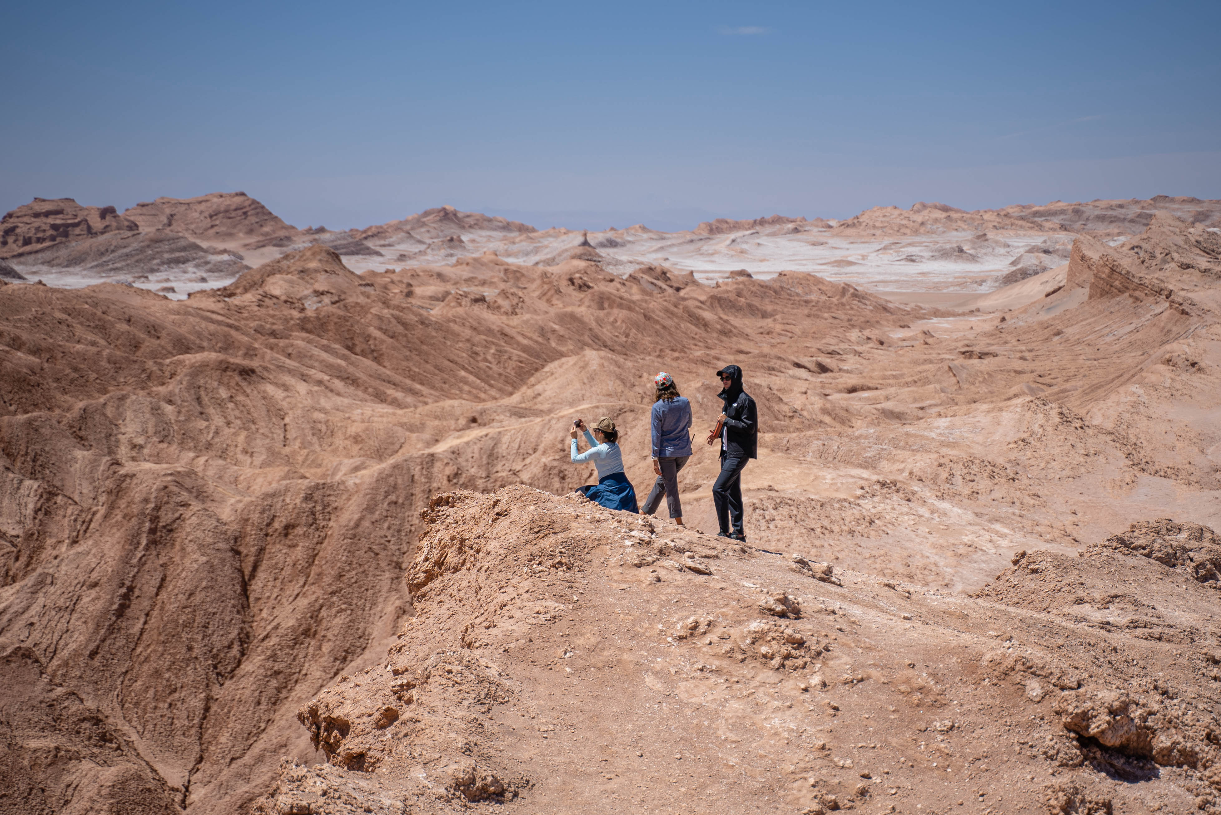 Small group hikes along a sandy ridge in a desert canyon, casting short shadows under bright sun high above the valley.