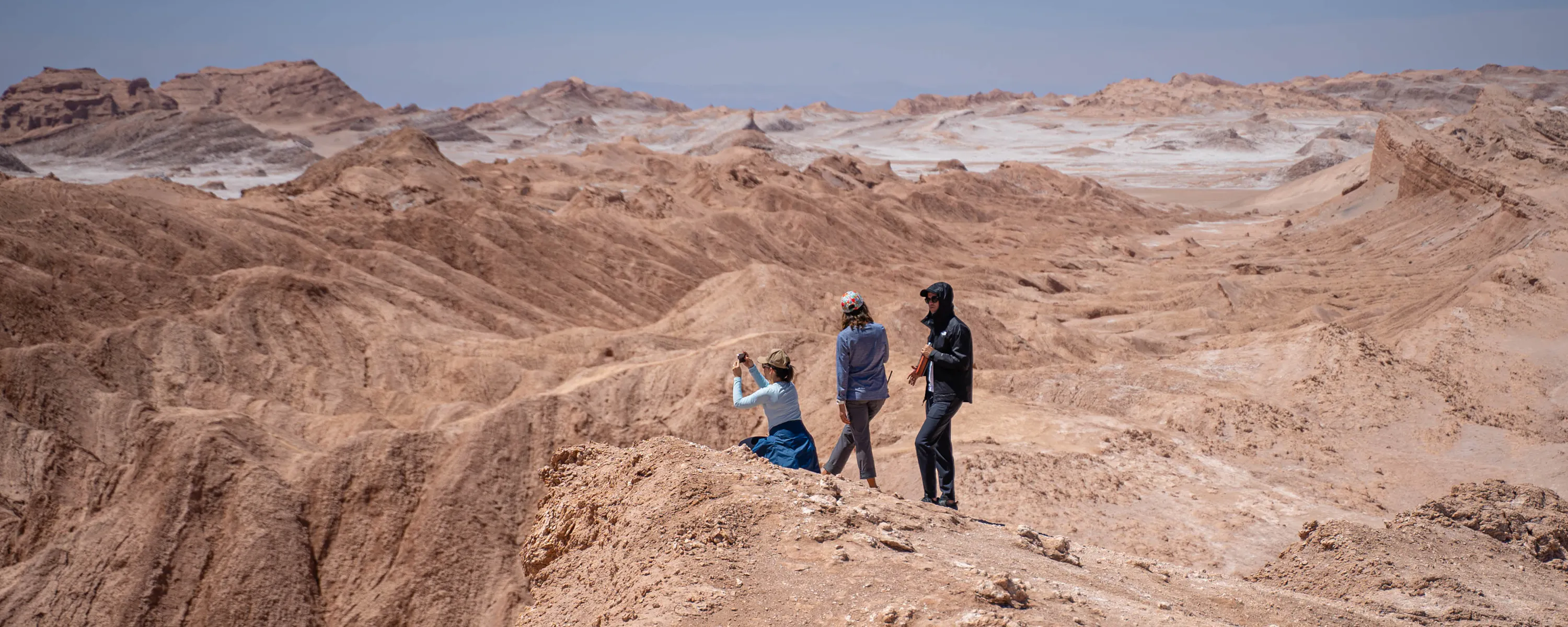 Small group hikes along a sandy ridge in a desert canyon, casting short shadows under bright sun high above the valley.