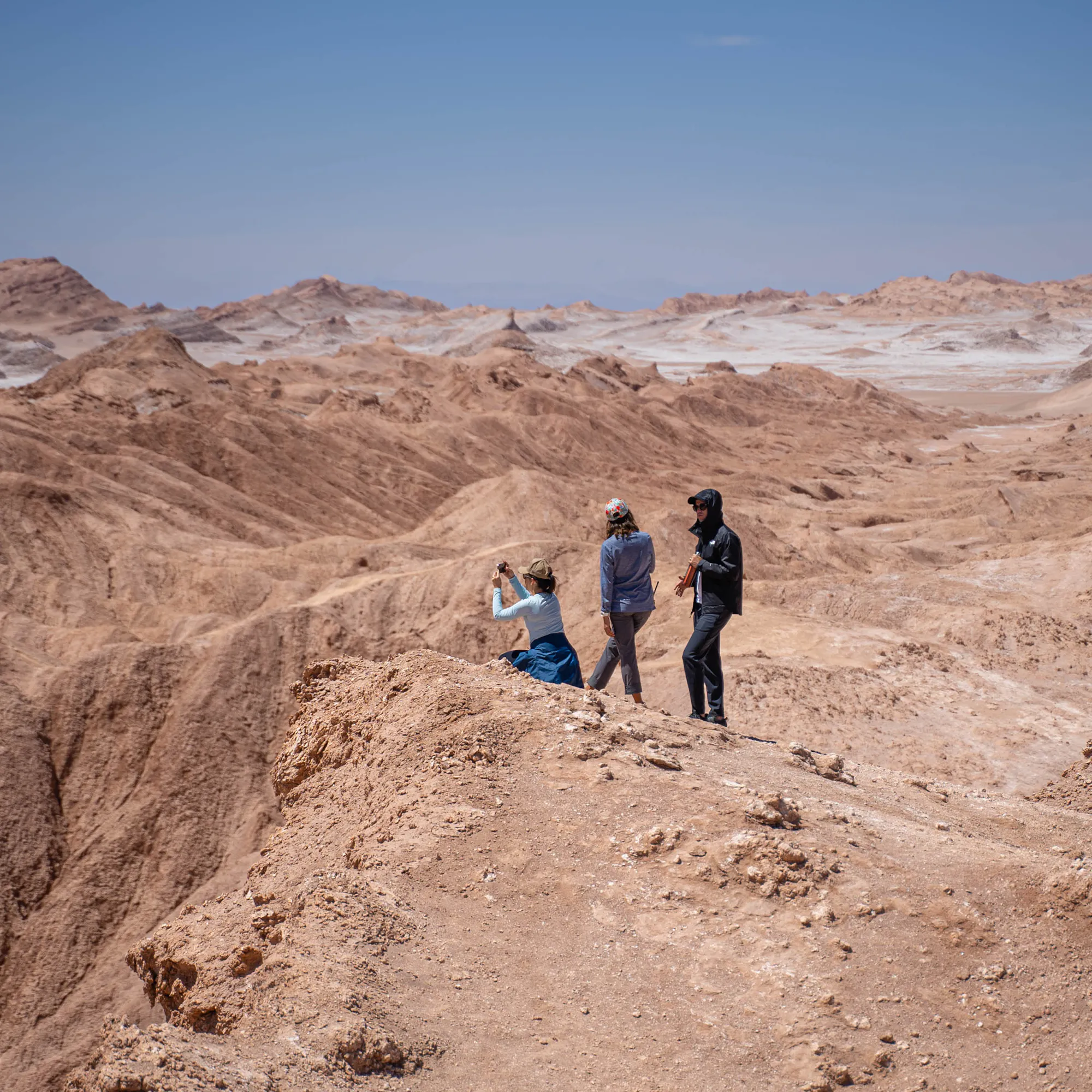 Small group hikes along a sandy ridge in a desert canyon, casting short shadows under bright sun high above the valley.