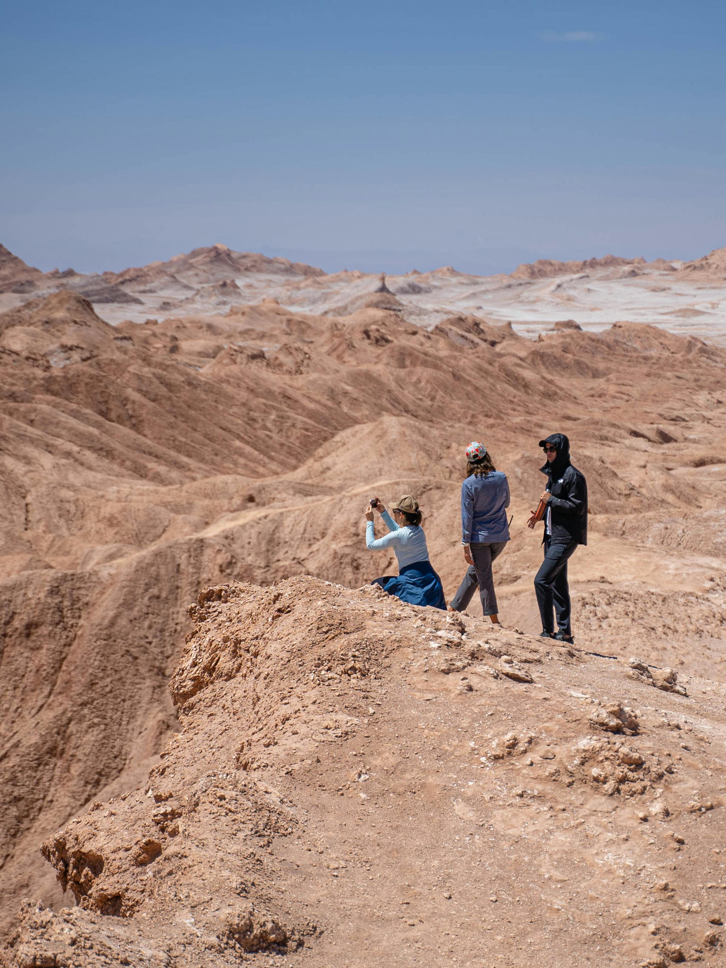 Small group hikes along a sandy ridge in a desert canyon, casting short shadows under bright sun high above the valley.