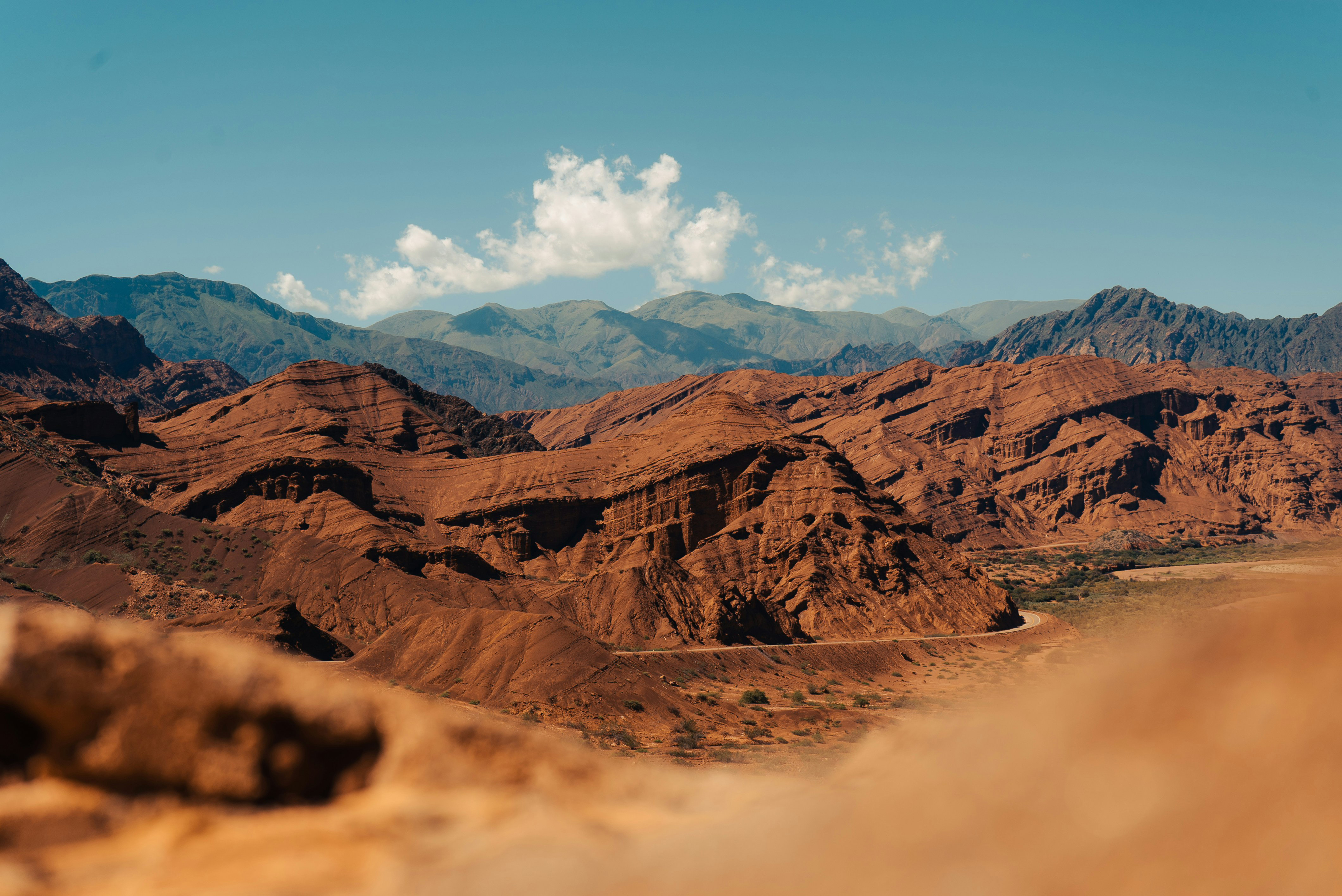 Red desert ridges stretch across a wide valley, with layered mountains and scattered clouds in the blue sky above them.