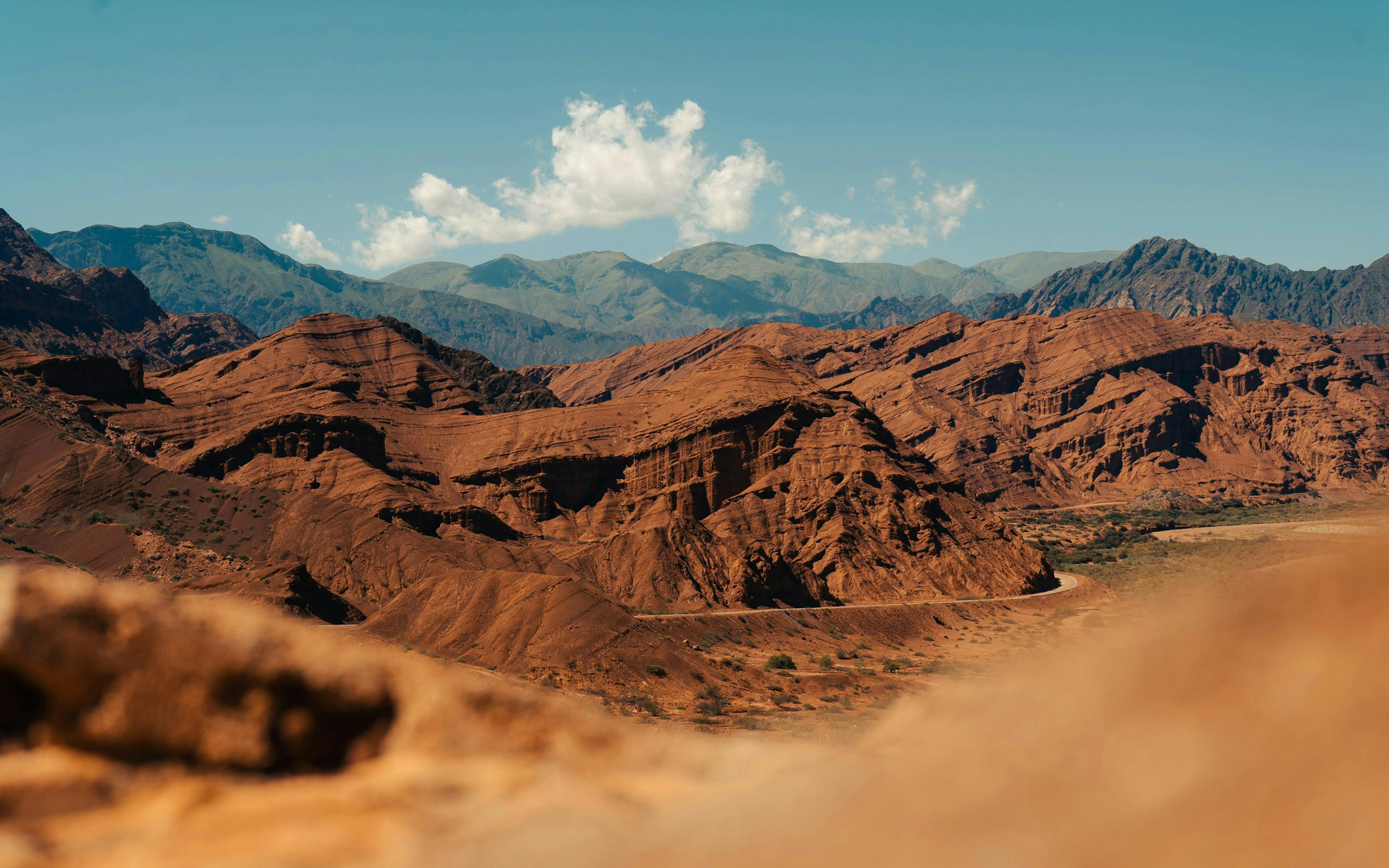 Red desert ridges stretch across a wide valley, with layered mountains and scattered clouds in the blue sky above them.