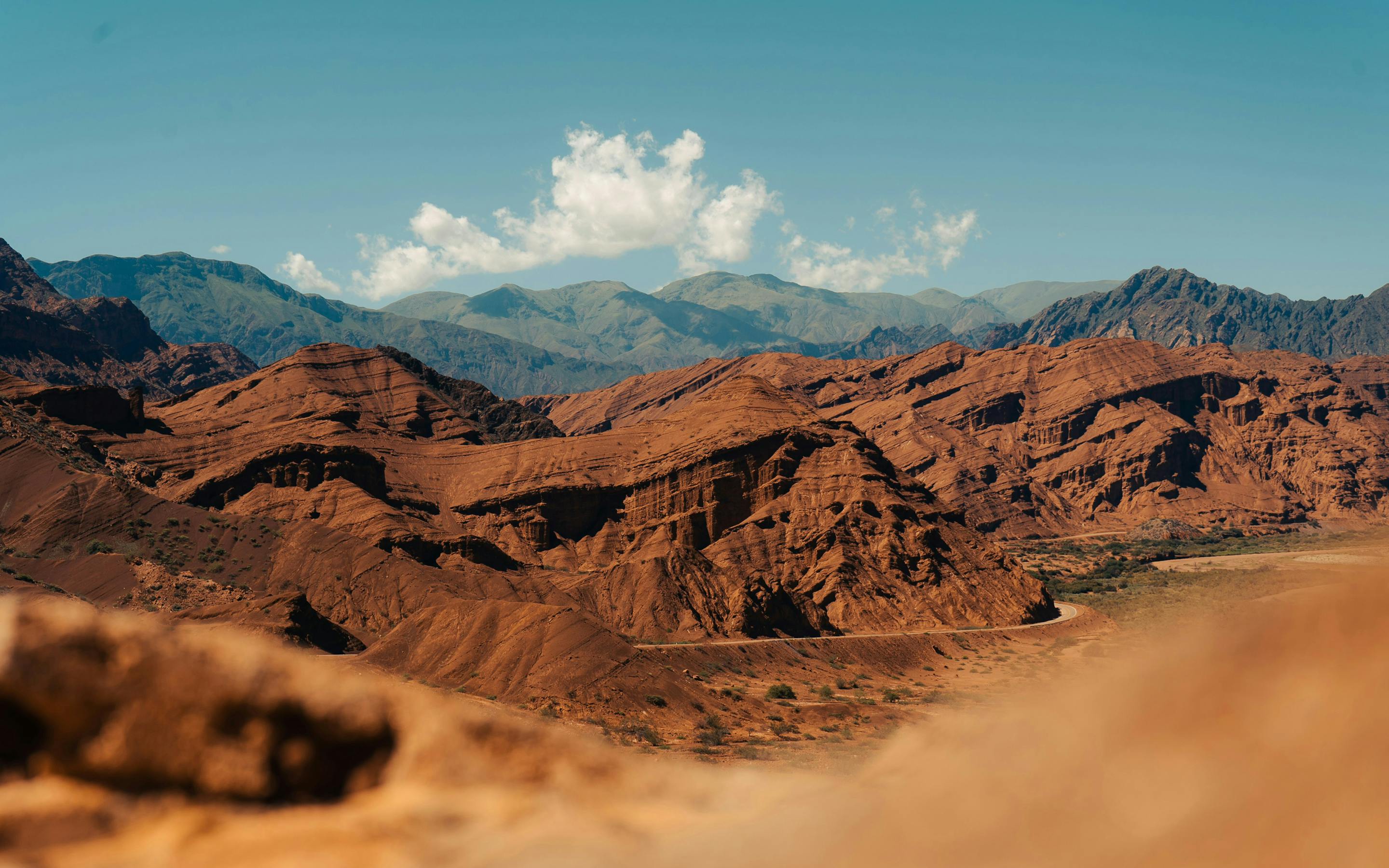Red desert ridges stretch across a wide valley, with layered mountains and scattered clouds in the blue sky above them.