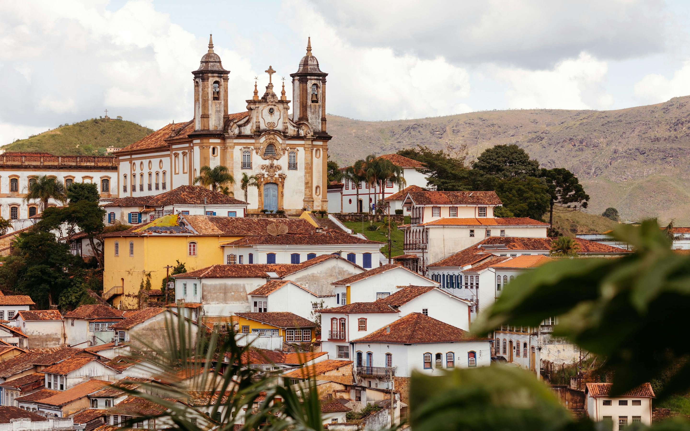 Colonial town spreads across a hillside, with twin church towers and colorful rooftops under a bright, cloudy sky in day light.