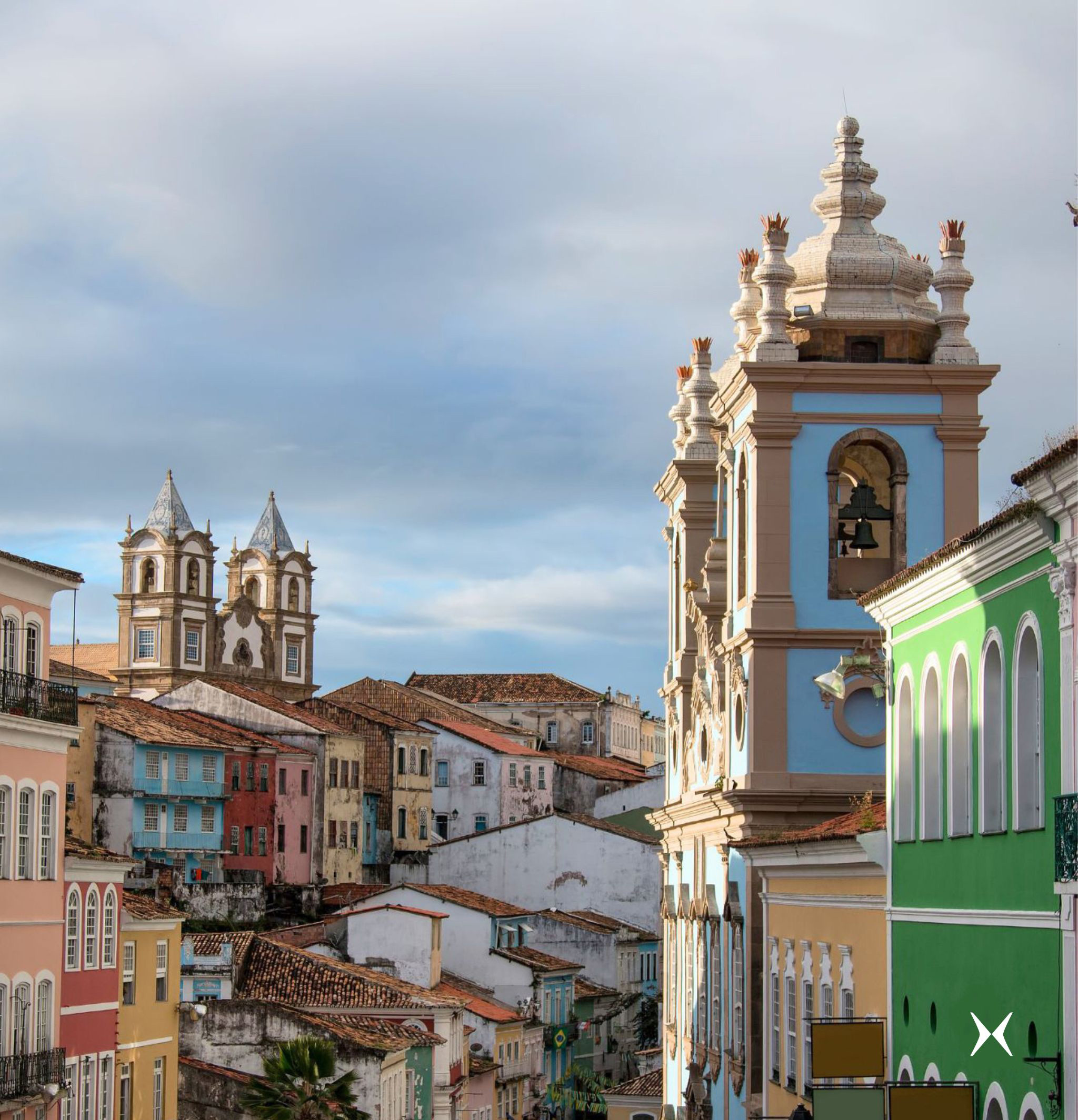 Historic church tower rises above colorful buildings, with tiled roofs and cloudy sky creating a layered city view below it.