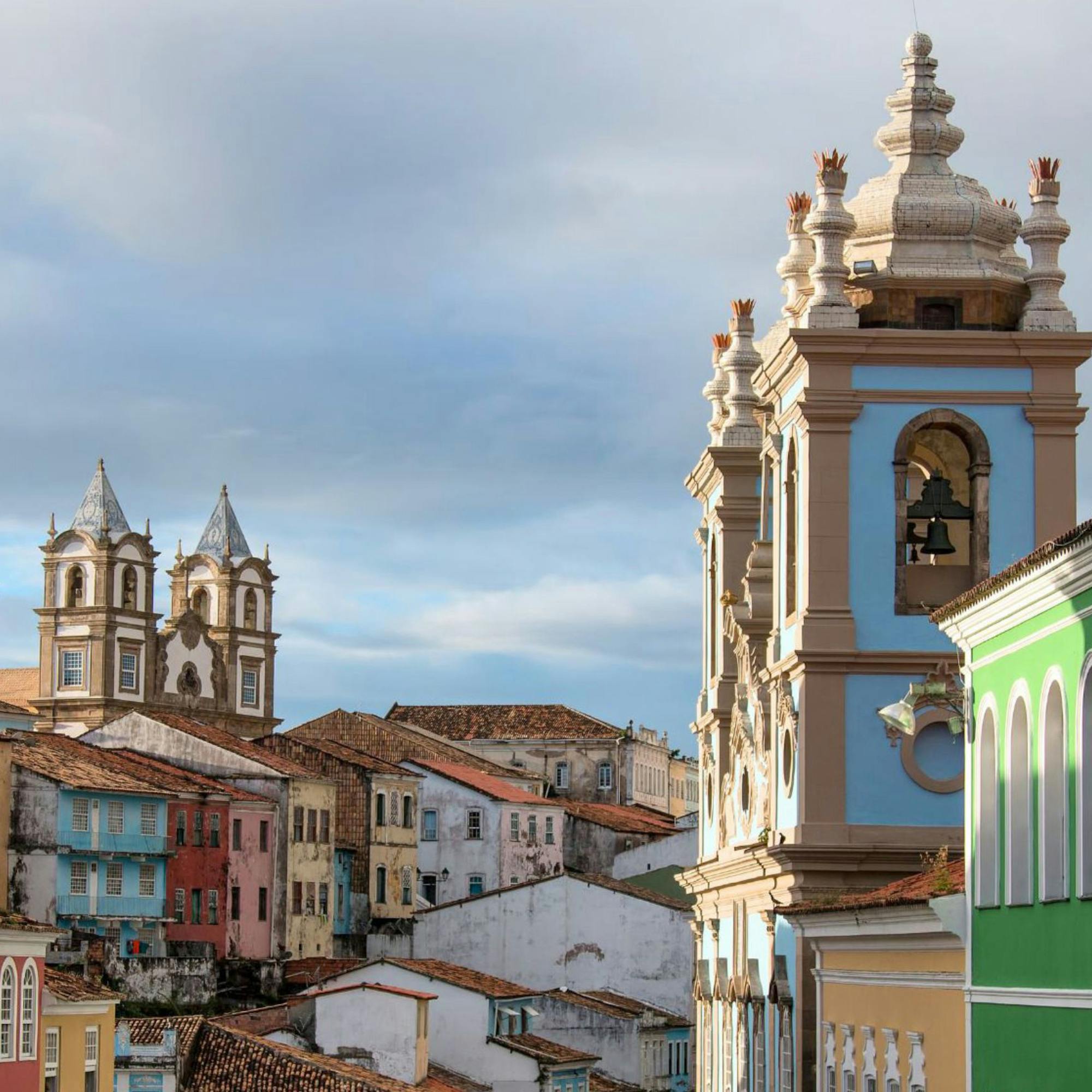 Historic church tower rises above colorful buildings, with tiled roofs and cloudy sky creating a layered city view below it.