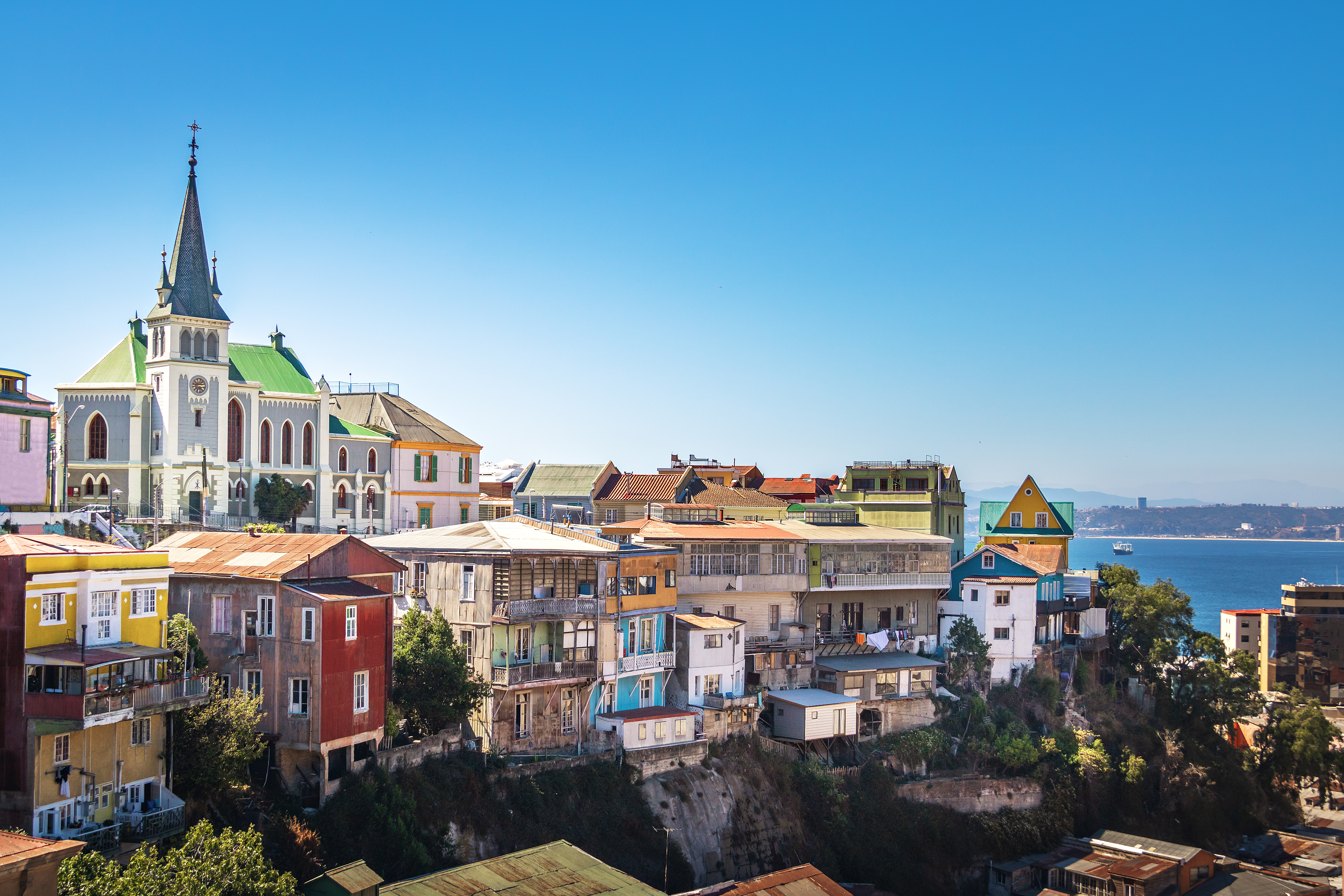 Colorful houses climb a steep hillside above the harbor, with a church spire and blue sky in view beyond them.