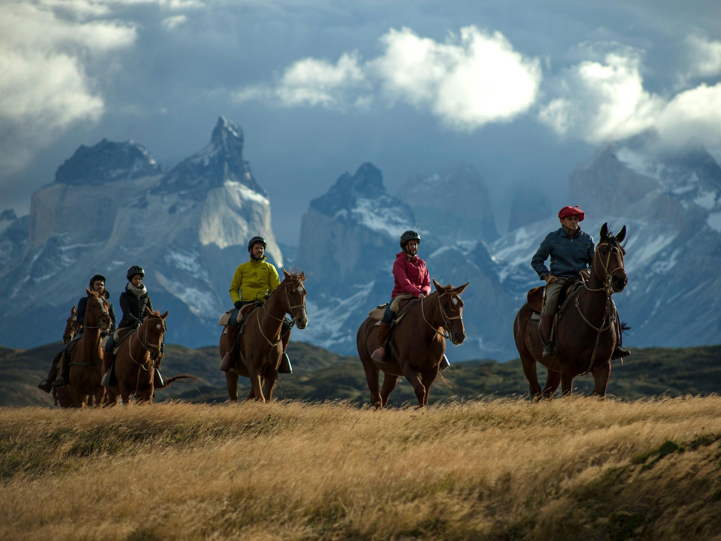 Group of riders on horseback crosses open grassland, with dark mountains and shifting clouds in the distance behind them.