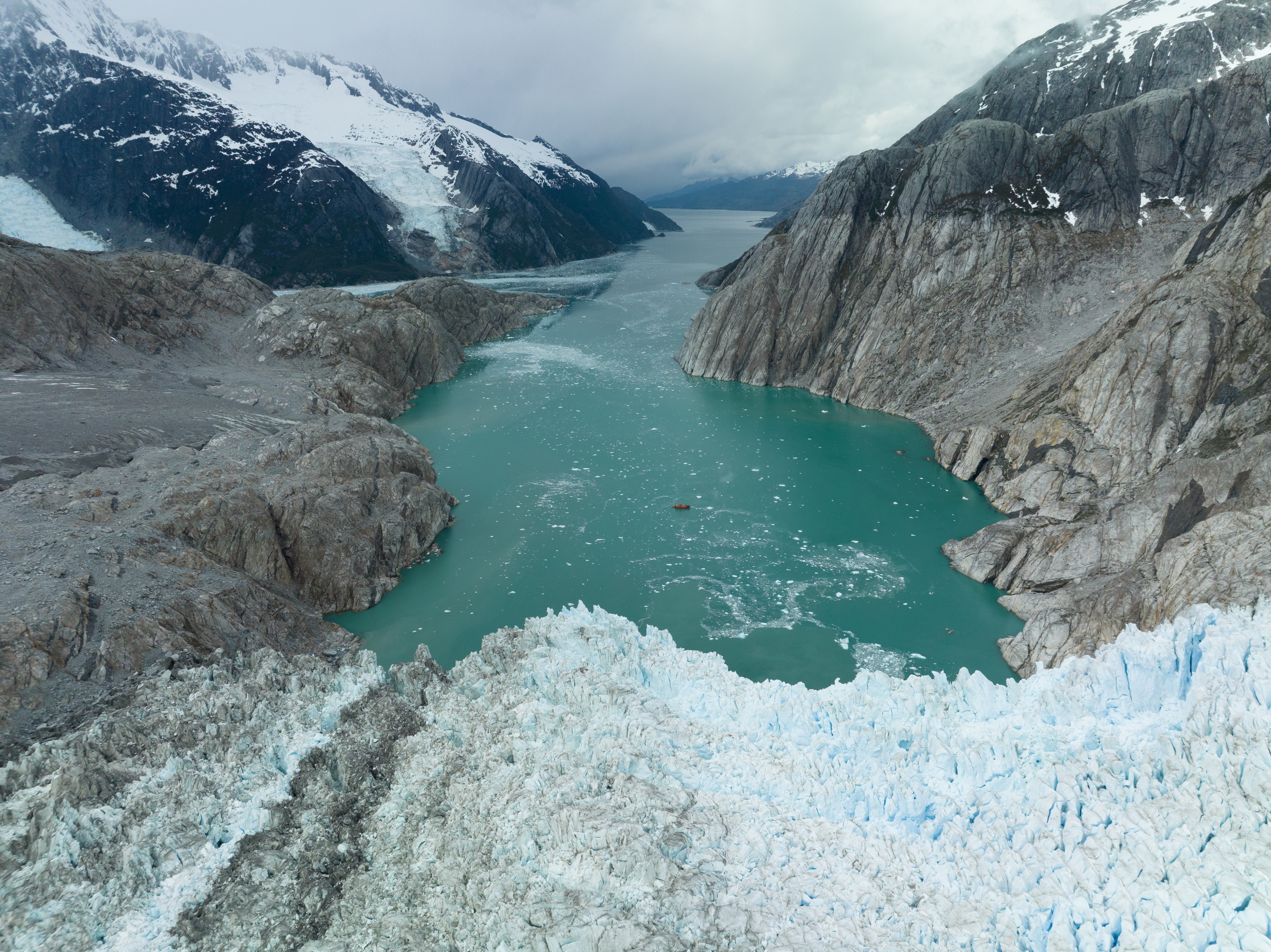 Turquoise glacial water meets cracked ice and rock, with dark mountains framing the frozen landscape from above.