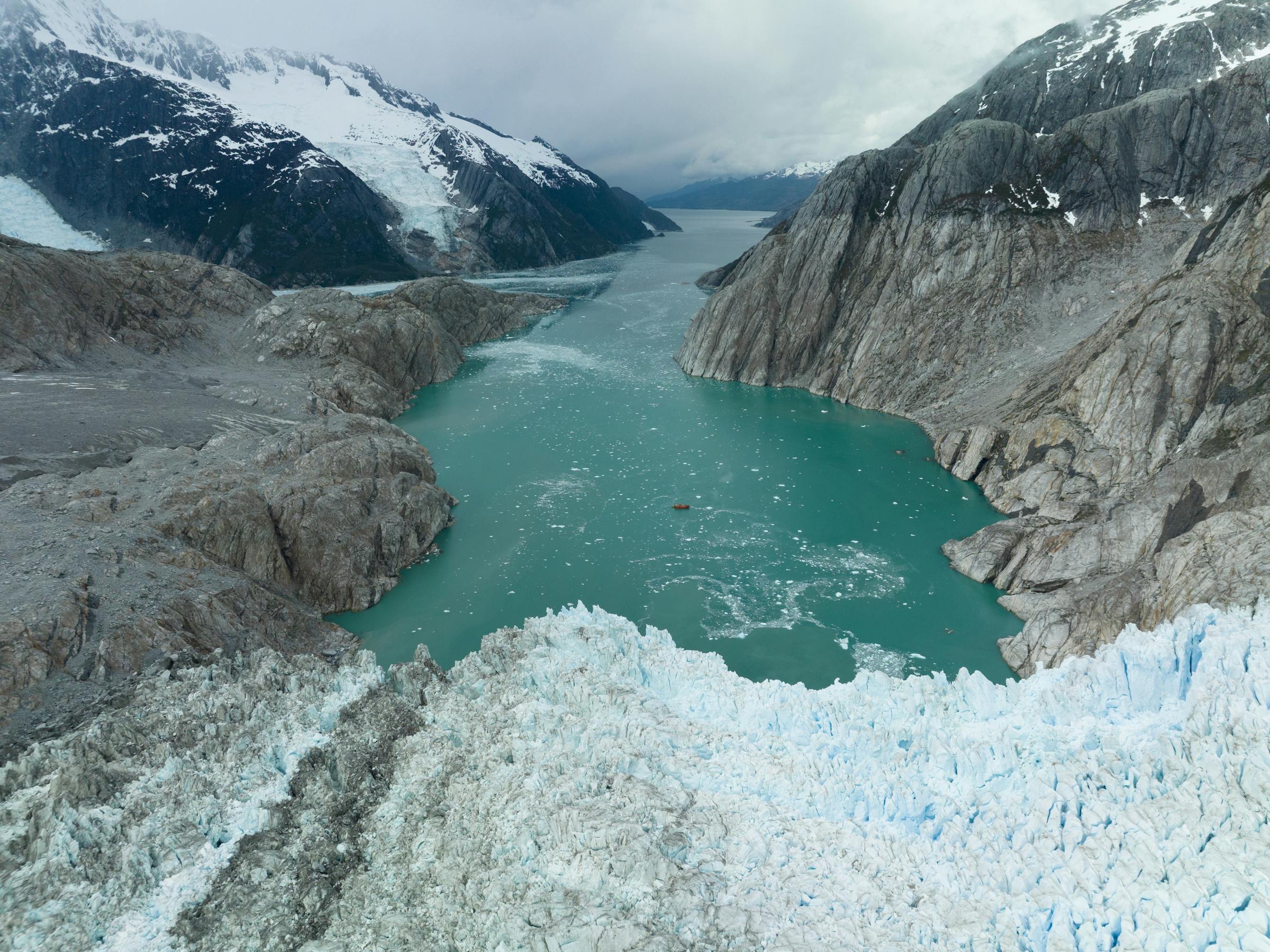 Turquoise glacial water meets cracked ice and rock, with dark mountains framing the frozen landscape from above.