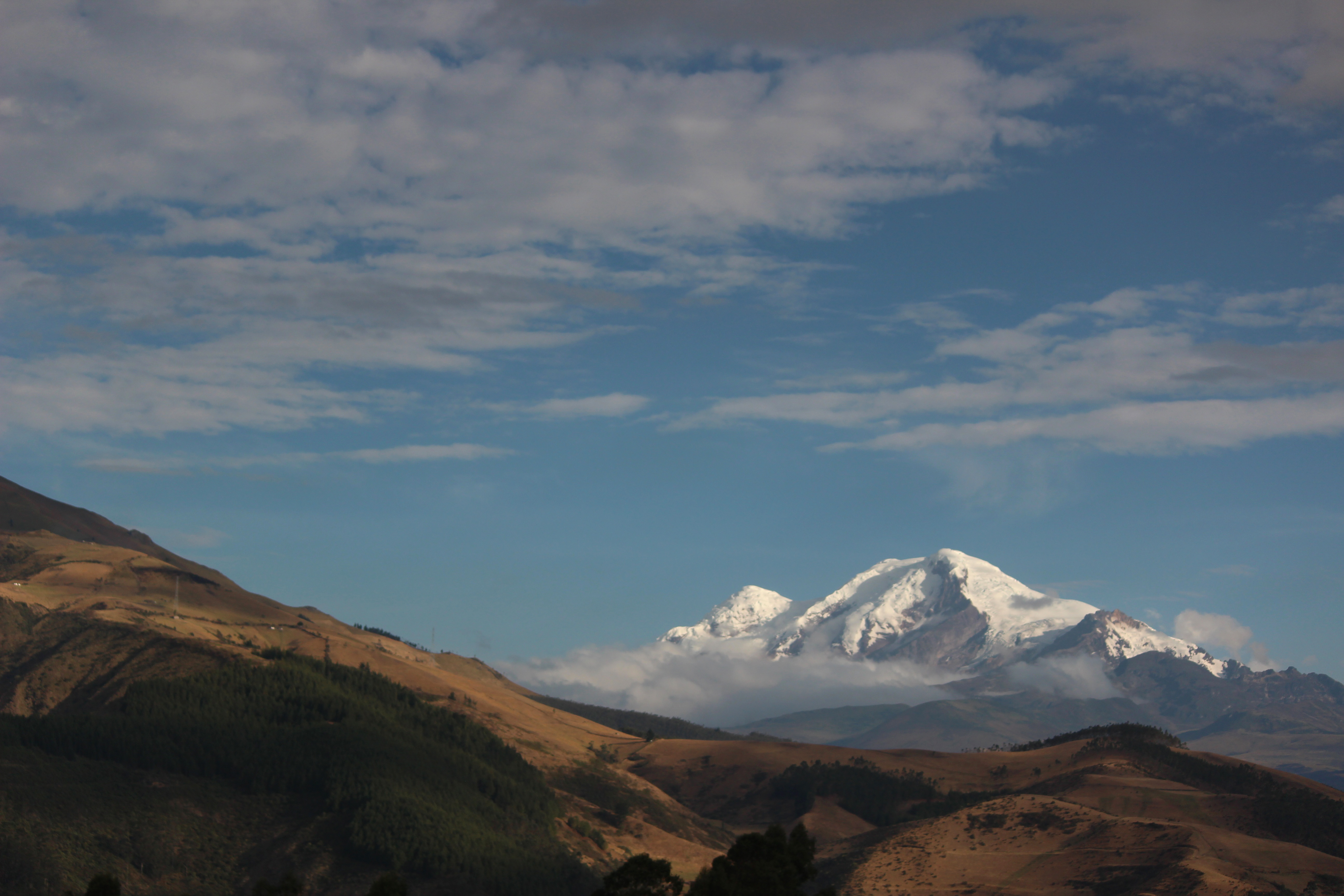 Golden hills lead to a snowcapped volcano beneath a broad blue sky, with scattered clouds over the valley.