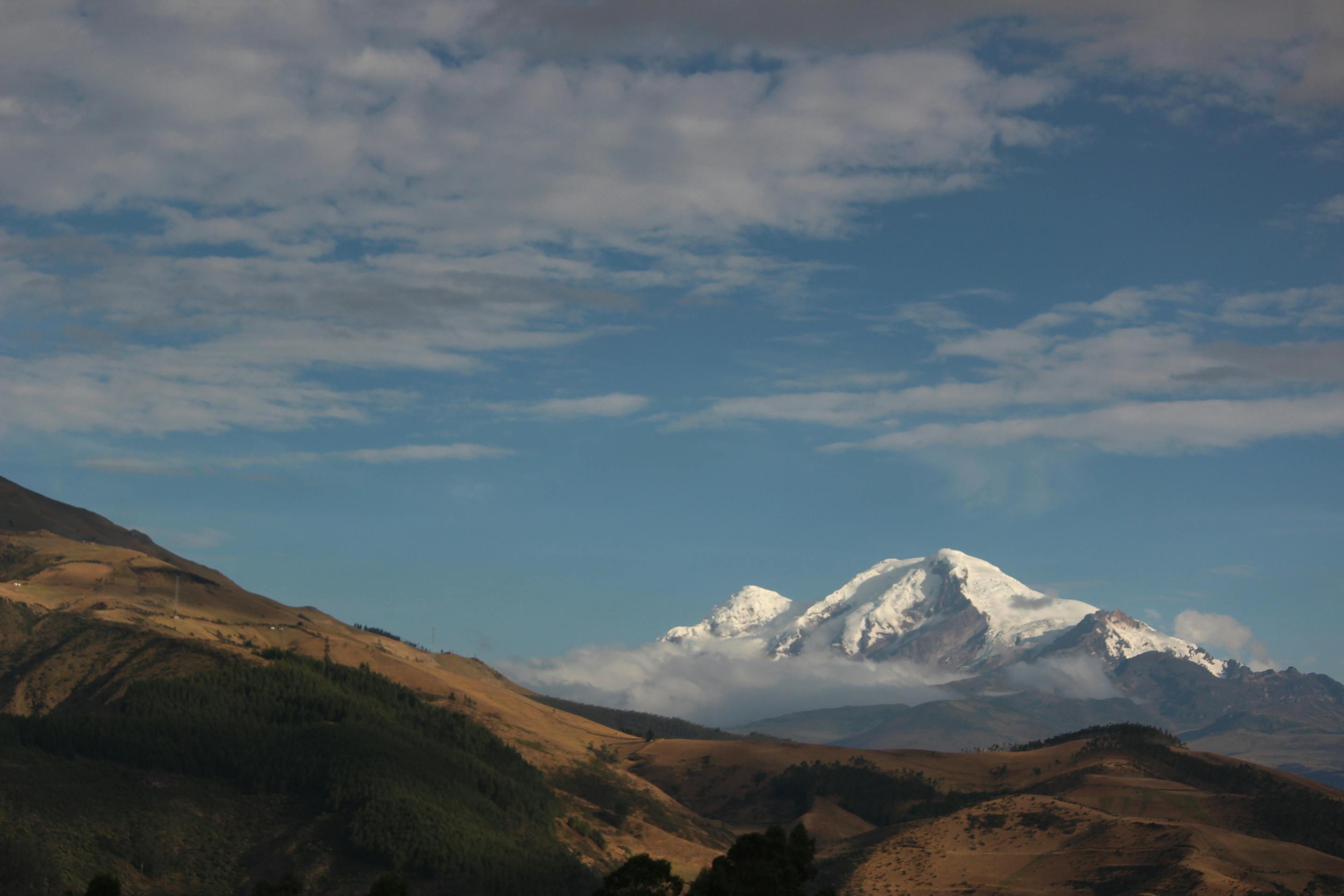 Golden hills lead to a snowcapped volcano beneath a broad blue sky, with scattered clouds over the valley.